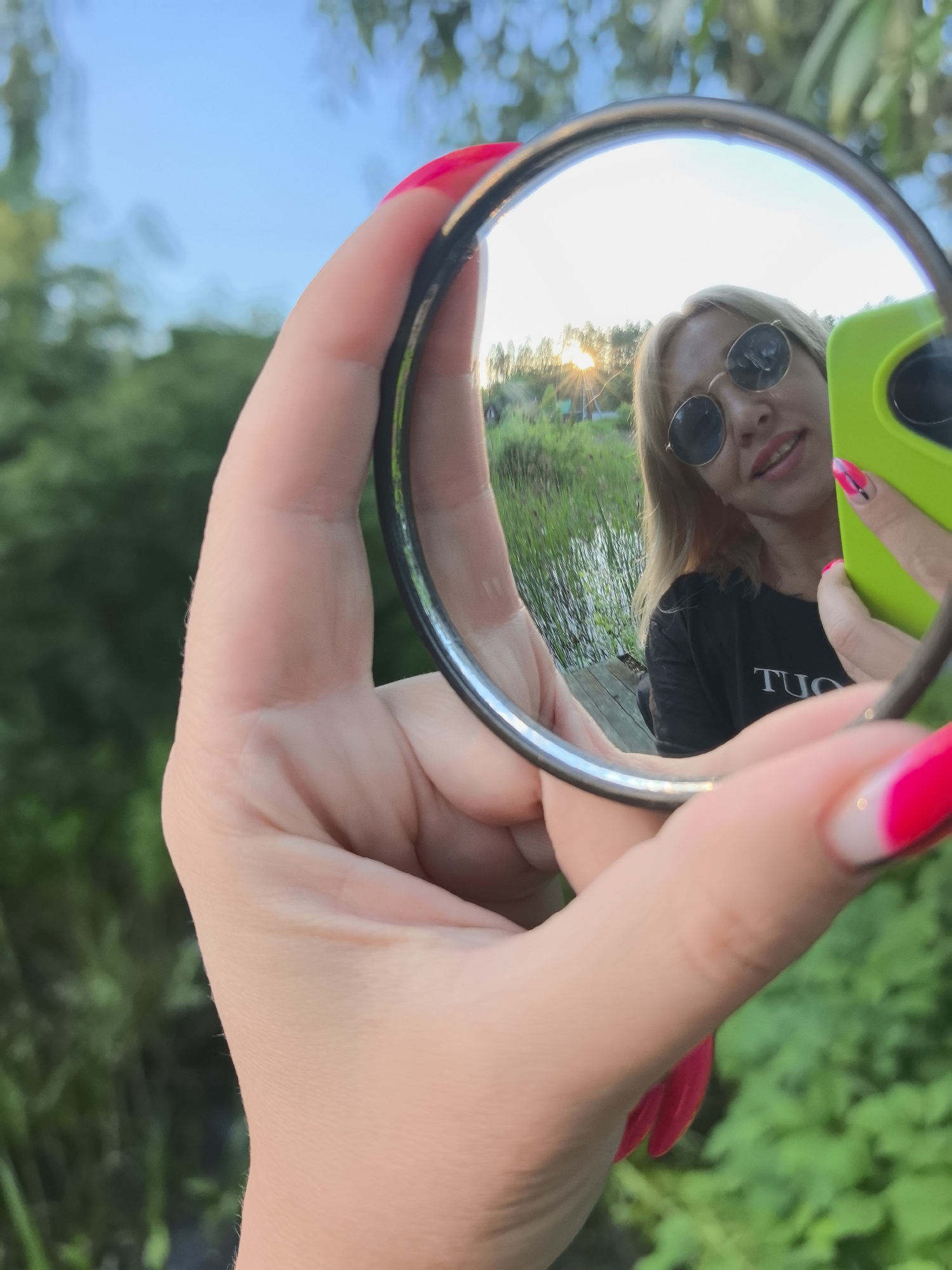A person is holding a round mirror in their right hand, reflecting a portion of themselves and the surrounding environment. The person in the reflection is a woman with blonde hair, wearing round sunglasses and a black t-shirt with white text that appears to say "TUO". She is smiling and holding a bright green smartphone in her left hand. The reflection also shows a body of water with reeds, a wooden dock, and trees in the background. The sun is setting, casting a warm glow and sun flares. The foreground of the image shows the hand holding the mirror, with bright pink manicured nails. The overall scene suggests an outdoor setting, possibly near a lake or pond, during sunset. The location is not specified in the image.