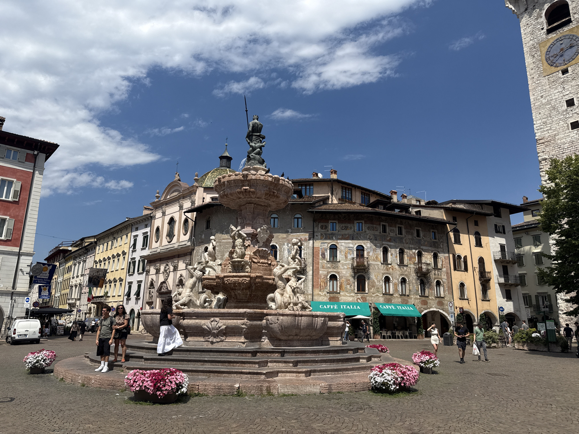 The image captures a bright, sunny day in Piazza del Duomo in Trento, Italy. A large, ornate fountain with multiple tiers and sculpted figures stands prominently in the center of the square. The fountain is adorned with statues of mythical creatures and figures, with a central statue of a man holding a spear, possibly Neptune. Surrounding the fountain, people are seen walking, standing, and interacting. Two individuals are posing near the fountain, one man in a green t-shirt and black shorts, and a woman in a leopard-print top and black skirt. Another woman in a white dress is walking away from the fountain.
The square is paved with cobblestones. Large planters filled with vibrant pink and white flowers are placed around the base of the fountain, adding color to the scene. In the background, a row of historic buildings lines the square. These buildings exhibit traditional Italian architecture with colorful facades, arched windows, and balconies. Two cafes with green awnings, labeled "CAFFÈ ITALIA," are visible, with tables and chairs set up outside. A white van is parked on the left side of the square, near a building with a red facade. Further to the right, a tall, stone clock tower stands against the blue sky, a prominent landmark of Trento. The sky is a clear blue with scattered white clouds. The overall atmosphere is lively and picturesque, showcasing a typical day in a European city square.