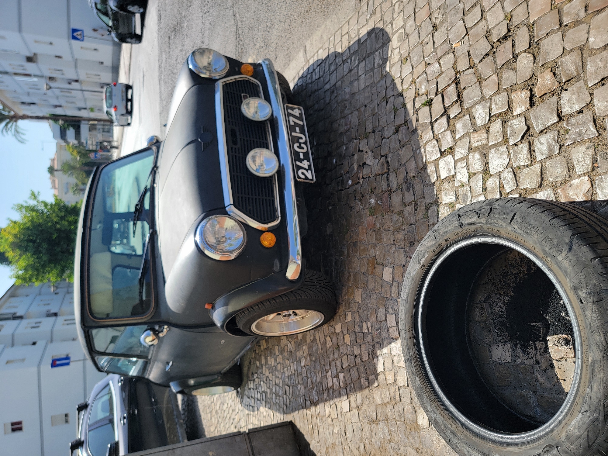 A matte black classic Mini Cooper is parked on a cobblestone street in Quarteira, Portugal. The car's front is facing the viewer, and it has a chrome grille, two large headlights, two smaller fog lights, and a license plate reading "24-CJ-74". In the foreground, a deflated tire lies on the cobblestones, partially obscuring the lower portion of the Mini Cooper. In the background, there are white buildings, trees, and other vehicles. One vehicle in the background appears to be a white sedan. The scene is illuminated by bright sunlight, casting shadows on the street.