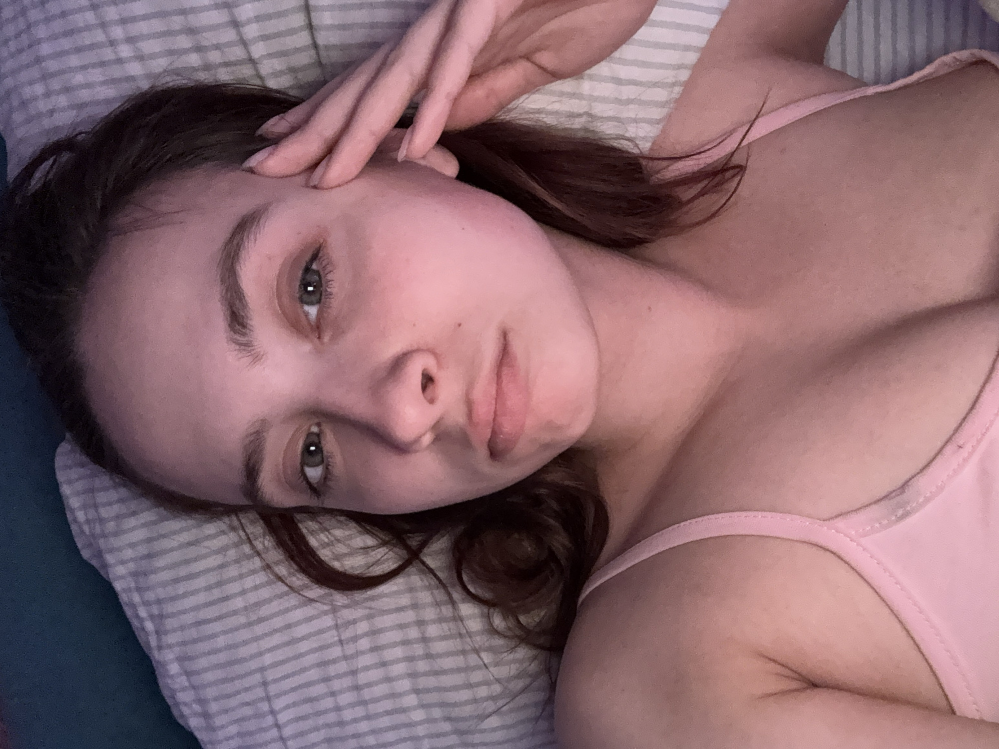 A young woman with fair skin, auburn hair, and green eyes lies on her back on a pillow with grey and white stripes. She wears a pink tank top. Her right hand rests on her forehead, fingers slightly spread. The background includes a teal-colored surface beneath the pillow. The location is unknown, including whether it is in or near the city of Прикамский, Russia.