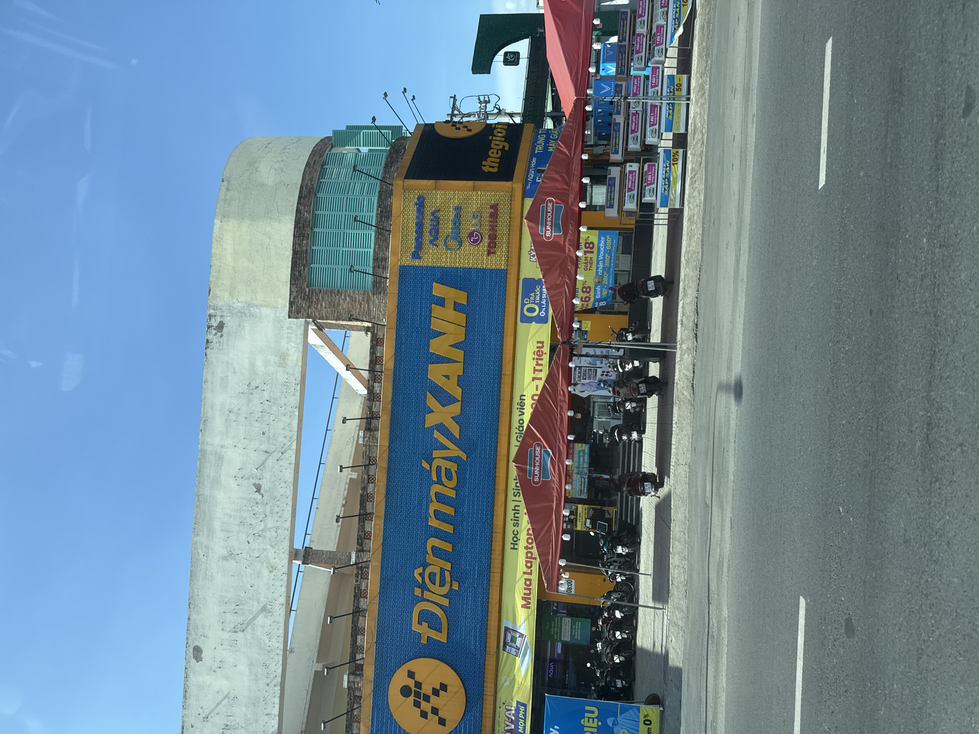 This is a horizontal, eye-level, medium shot of the exterior of a Điện Máy XANH electronics store on a sunny day in Thuận An, Vietnam. The store's blue and yellow facade is prominent, featuring a large "Điện máy XANH" logo and various brand names like Panasonic, Aqua, Midea, LG, and Toshiba. In front of the store, several red tents are set up, with motorcycles parked behind them. The tents appear to be displaying promotional items or merchandise. Below the tents, various sale signs are visible, advertising discounts and payment plans. A street with a white lane marker runs in the foreground, with a portion of a concrete sidewalk visible between the street and the store. The sky is a clear blue. There are no people clearly visible in the scene.