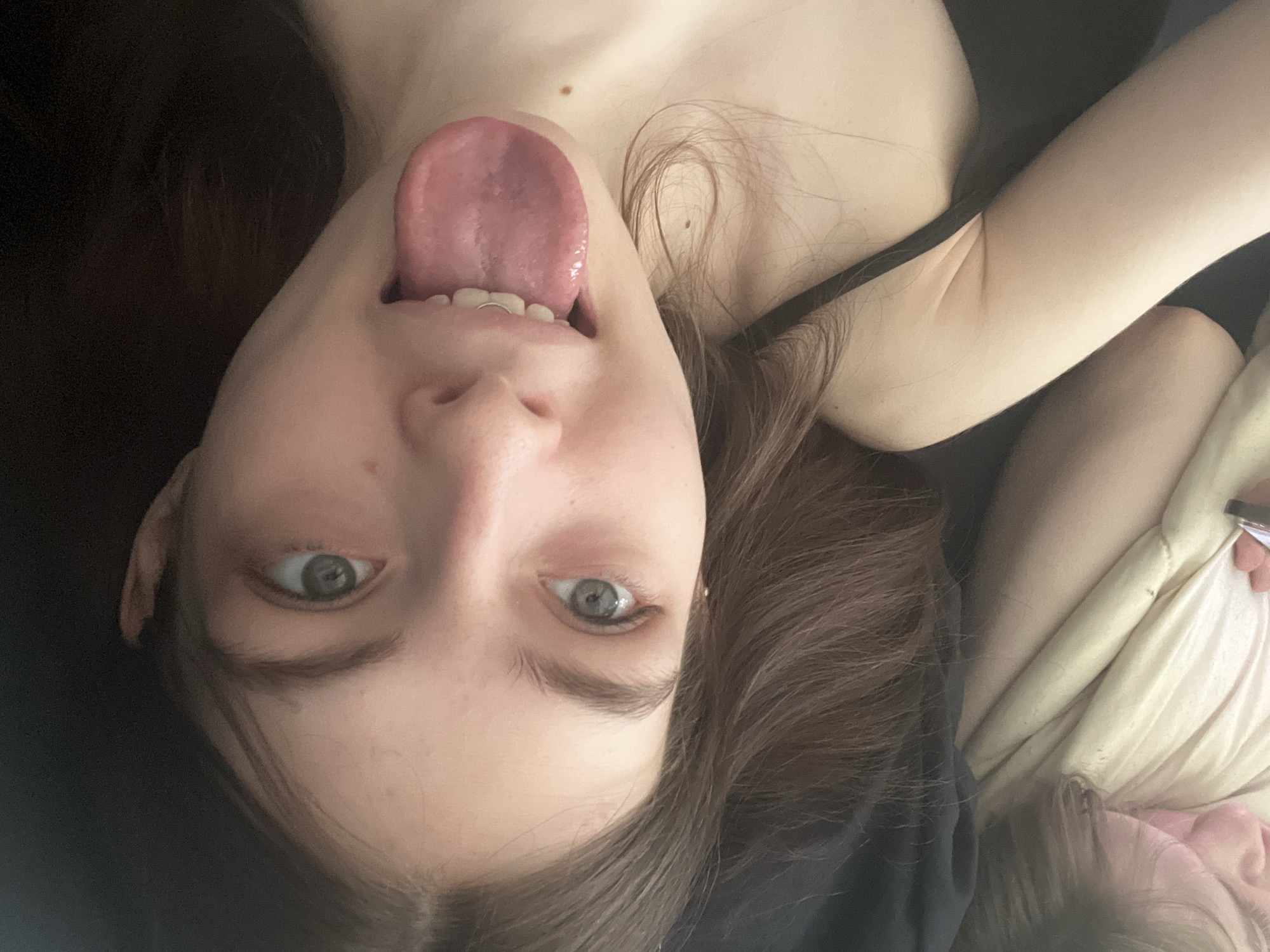 A young, fair-skinned person lies with their head upside down, tongue extended. The background is dark, suggesting an interior setting. The person has brown hair and light-colored eyes. The image appears to be taken at close range. No objects or details suggest the location is Saint Petersburg, Russia.