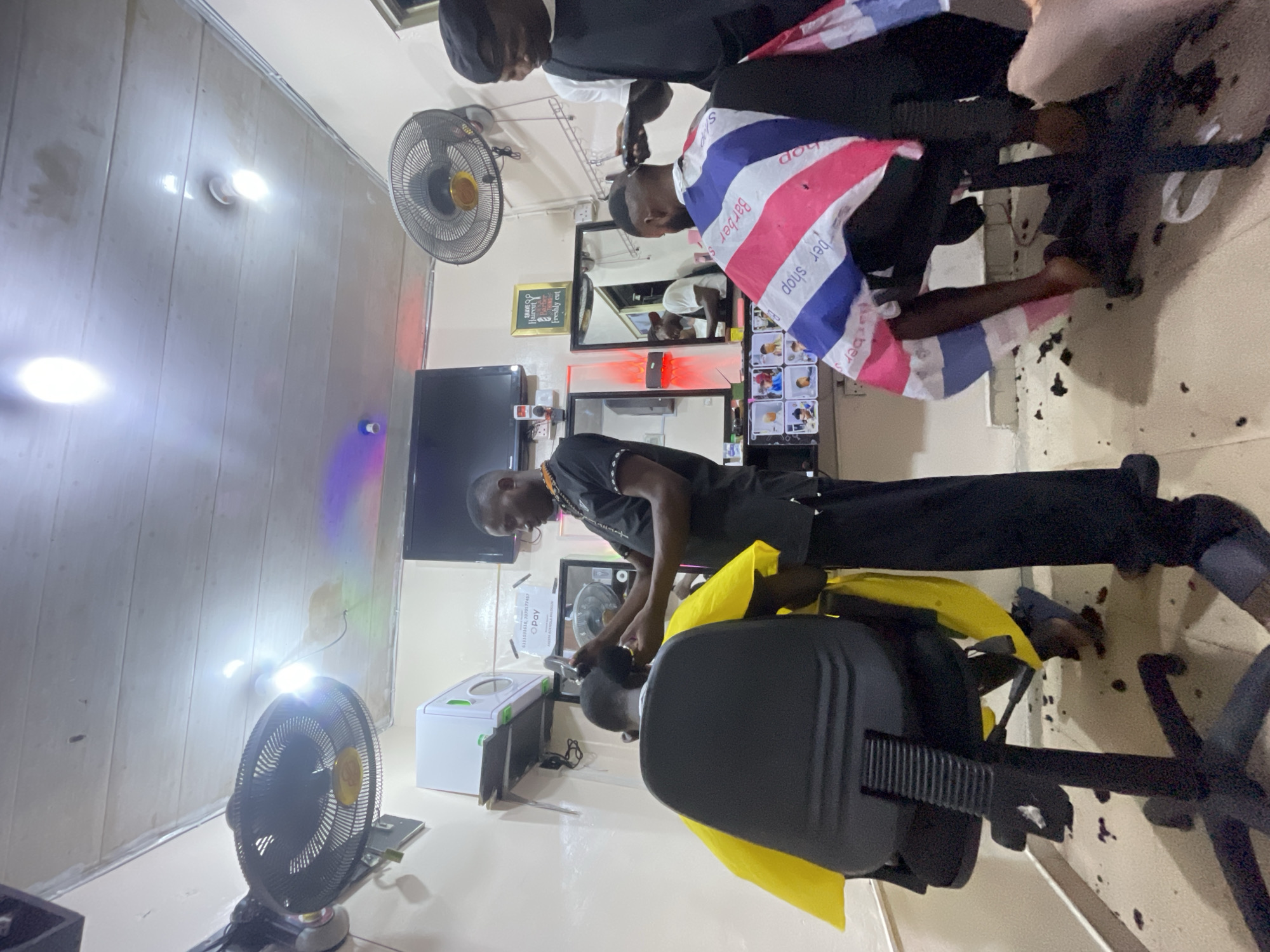 The image shows the interior of what appears to be a barbershop in Ado, Nigeria. Two barbers are actively cutting hair on two customers. One customer is draped in a red, white, and blue barber's cape. The second customer is draped in a yellow cape and seated in a black barber's chair. Various items are observed including two fans, a television, a mirror, and a container labeled "O-Pay". Hair clippings are visible on the floor. The environment seems to be well-lit.