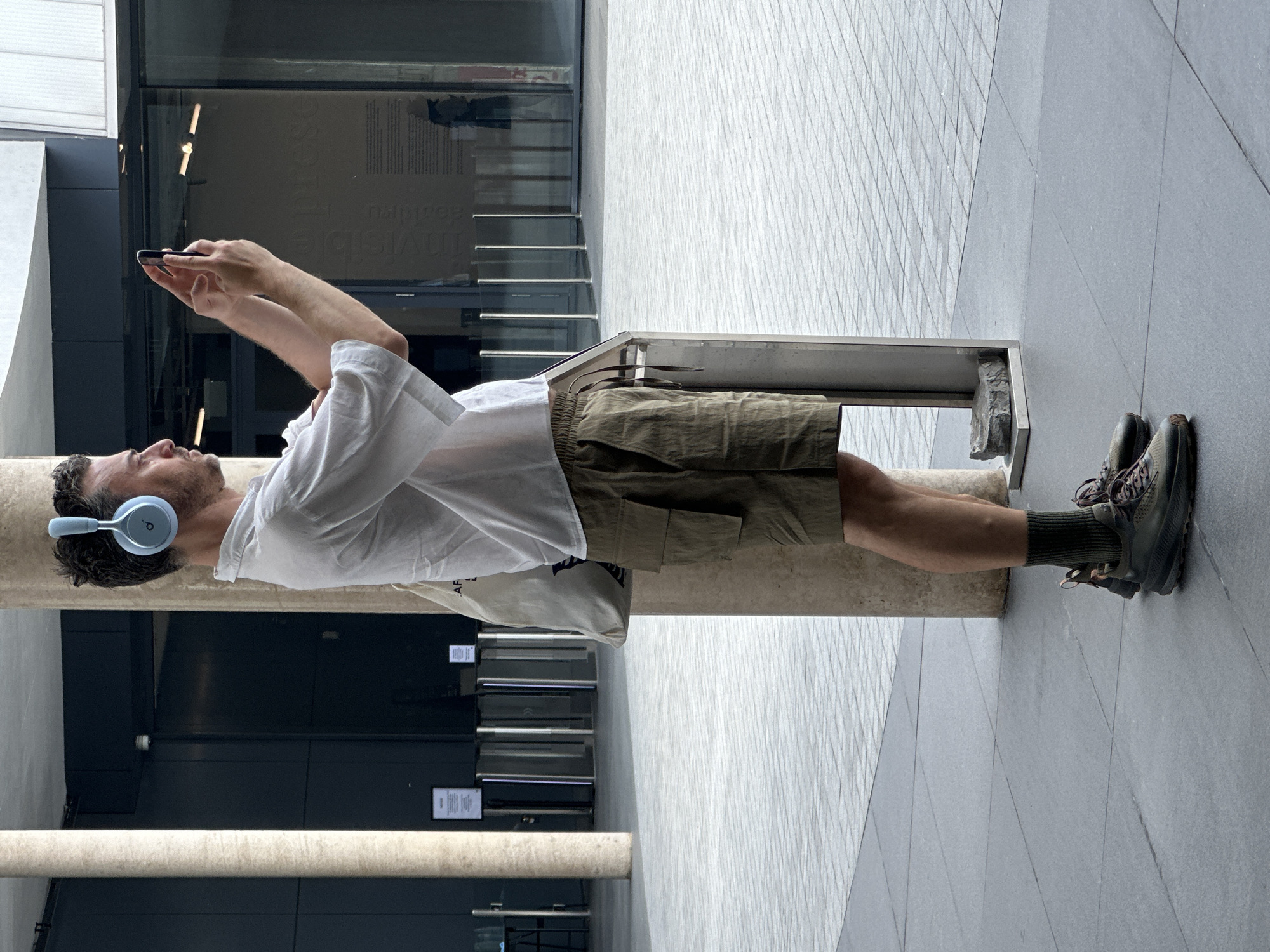 A man is lying on a concrete bench in an urban environment. He is wearing a white shirt, khaki shorts, and hiking shoes with dark socks. He has headphones on and is looking at his phone, which he holds in both hands. The bench is situated in front of a modern building with glass doors and a gray facade. The ground is paved with gray tiles. The image appears to be taken in Bangkok, Thailand, although specific details confirming the location are not visible.