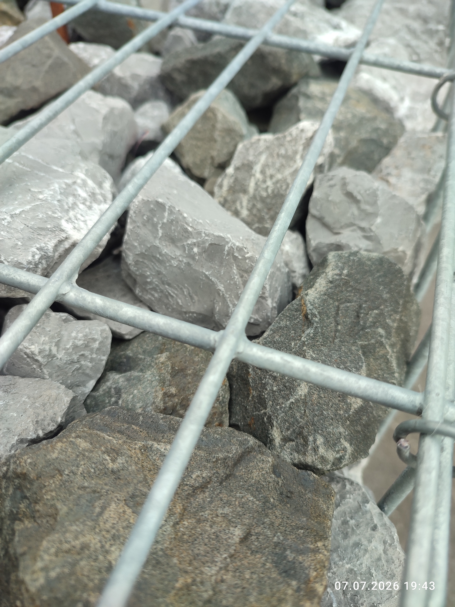 The image shows a close-up view of a gabion wall, which is a structure made of wire mesh filled with stones. The gabion is filled with various sizes of gray and dark gray rocks. In the foreground, two horizontal metal bars of the gabion cage run across the image, and a diagonal metal bar is also visible. Some of the rocks are sharp and angular, while others are more rounded. The image is taken outdoors, and the lighting suggests it is daytime. The bottom right corner of the image displays a timestamp: "07.07.2026 19:43". The scene is likely located in Immenstadt im Allgäu, Germany, based on the provided information. There are no people or discernible actions depicted in the image. The focus is on the texture and composition of the gabion and its contents.