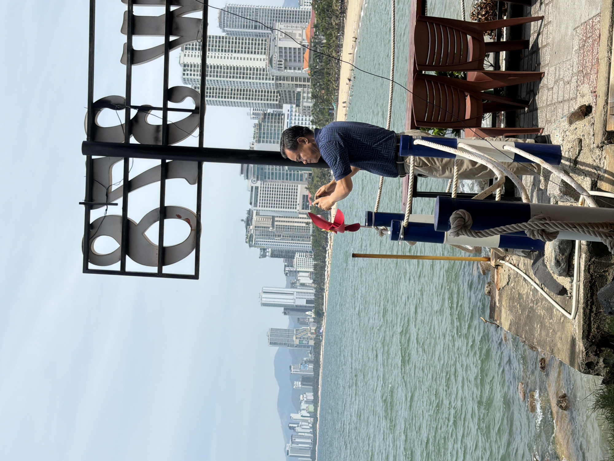 A man, possibly a tourist, stands on a pier in Nha Trang Ward, Vietnam, capturing a photo of a red pinwheel with his phone. He is dressed in a blue patterned shirt and light-colored pants. Behind him, the turquoise ocean stretches to a coastline dotted with palm trees and a dense collection of high-rise buildings. The sky is overcast, suggesting a possibly humid day. The pier is decorated with blue and white striped posts and thick rope railings. In the foreground, two brown plastic chairs are positioned near a table.