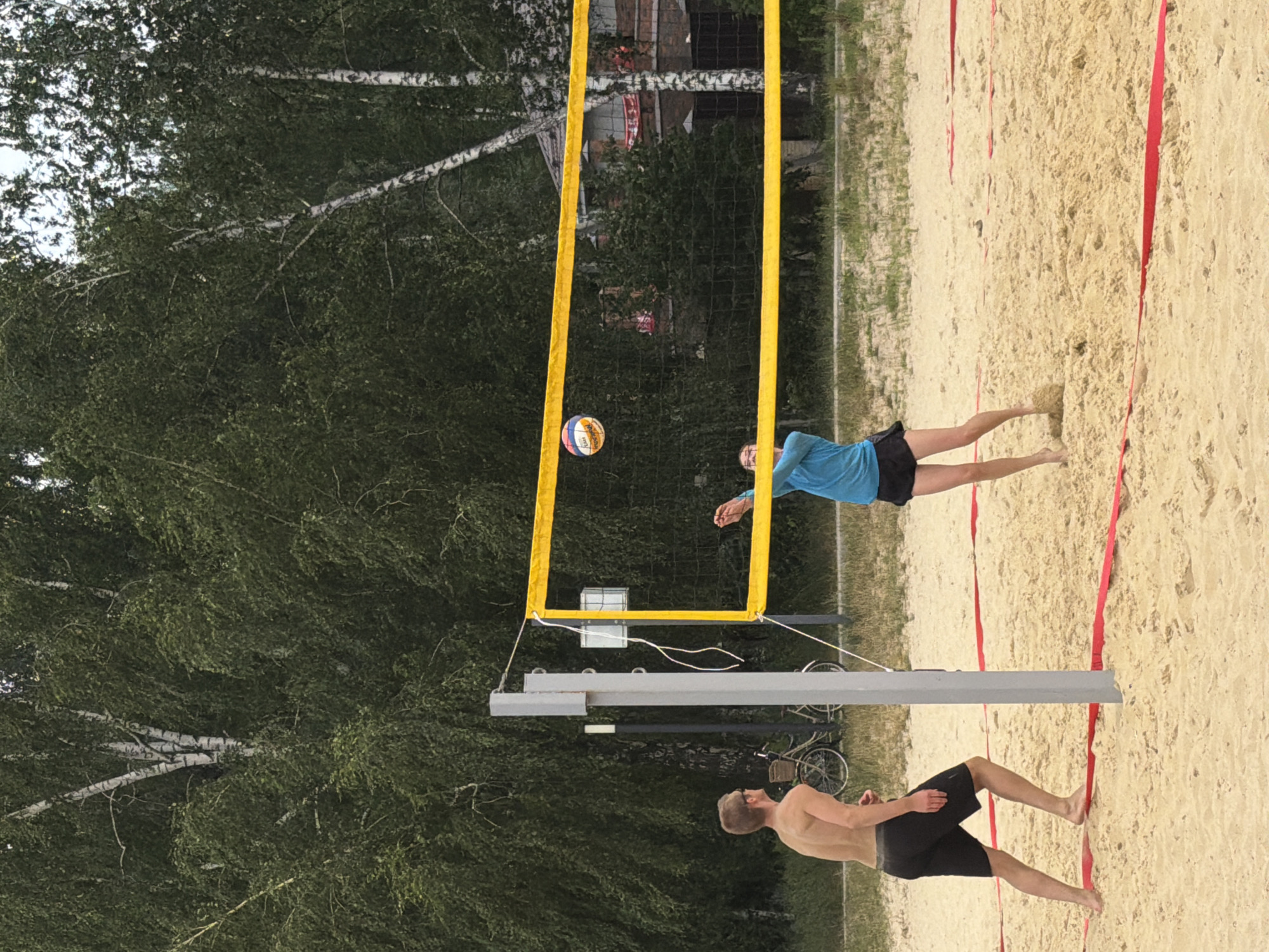 In Malakhovka, Russia, two individuals are playing beach volleyball on a sand court surrounded by trees. On the left, a shirtless male with dark shorts is positioned to receive a serve, facing the net. On the right, a female wearing a blue shirt and dark shorts is in the process of hitting a volleyball over the net. The volleyball is visible in mid-air, above and to the left of the female player. A yellow volleyball net with a yellow border stretches across the court, supported by a gray pole on the left. In the background, trees with white bark and green foliage dominate the scene. Two bicycles are visible parked on the grass behind the volleyball court. The ground is covered with sand, with red lines indicating the boundaries of the court.