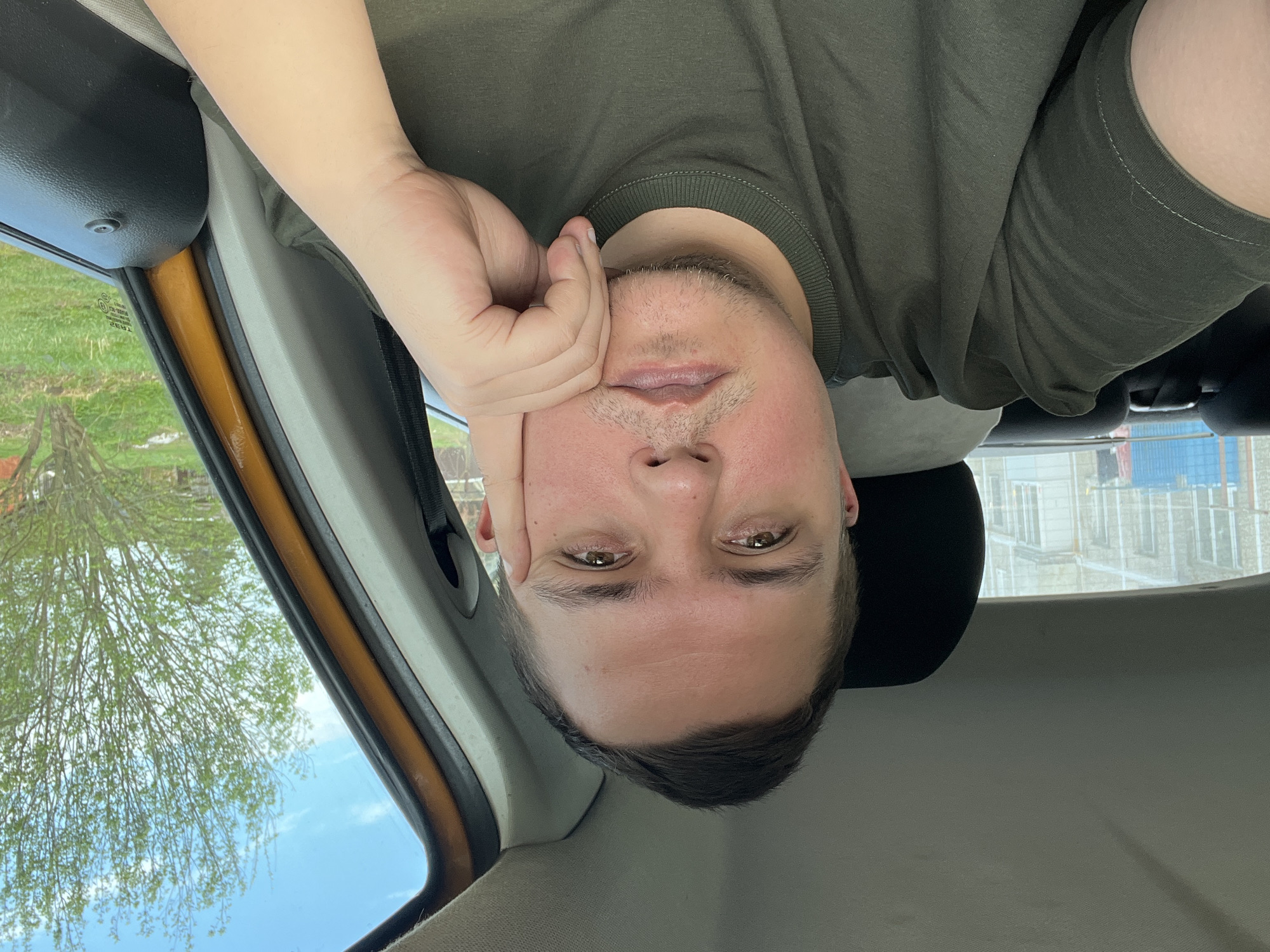 A selfie taken from inside a car in Razdolye, Russia, shows a fair-skinned man wearing a green t-shirt and resting his head on his hand. He looks directly at the camera. Outside the car window, trees are visible with new leaves indicating spring. A building under construction is also visible outside the passenger side window. The car’s interior is visible, including the door frame and a headrest.