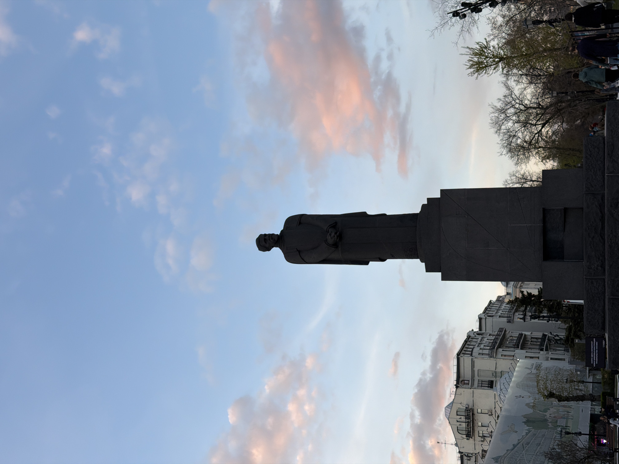 The image depicts a bronze statue of Alexander Griboyedov in Moscow, Russia, taken against a background of a sky with pinkish clouds. The statue stands atop a dark stone pedestal, with buildings visible in the distance. The statue has a slight bend with his head tilted to the side. A few people are visible in the background, partially obscured by trees. A mural is partially visible below the buildings on the right.