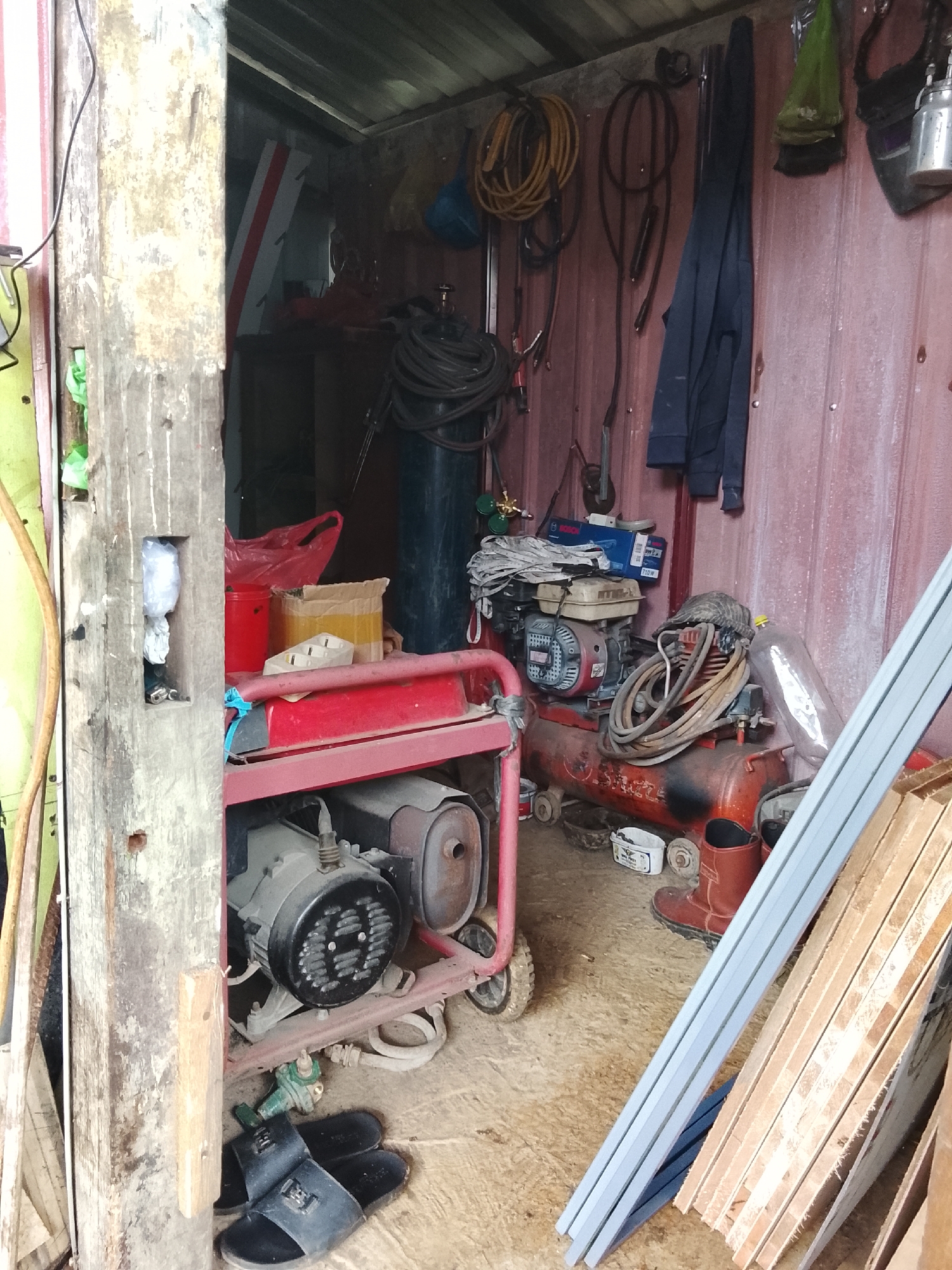 The image depicts the interior of a cluttered storage shed. A red generator with a black engine sits prominently in the foreground, positioned on a red frame with wheels. To the right of the generator is an orange air compressor, also on wheels, with various hoses coiled around it. Several pairs of black flip-flops are scattered on the floor in front of the generator. In the background, a large blue gas cylinder is visible, along with a dark-colored jacket hanging on the wall. The walls and ceiling of the shed appear to be made of corrugated metal. Several items are hung on the walls, including coiled hoses and bags. Stacked lumber is present in the right foreground. The overall impression is one of a functional, albeit disorganized, workspace or storage area. The location in Mee, Indonesia, is not discernible from the visual evidence.