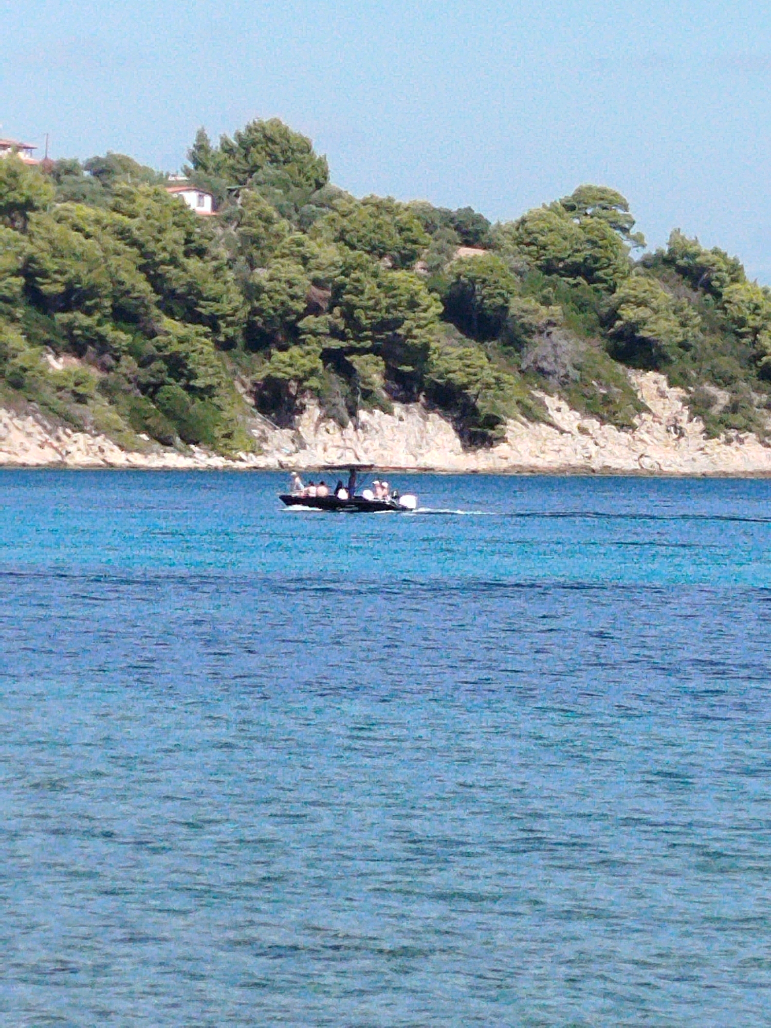 A small motorboat carrying several people navigates a calm, clear blue sea. The boat has a dark canopy providing shade for some of the passengers. In the background, a rocky, tree-covered hillside slopes down to the water. A few white buildings, possibly houses, are visible nestled amongst the trees on the hillside. The sky is a pale blue with no clouds. The scene suggests a serene summer day in a coastal location.