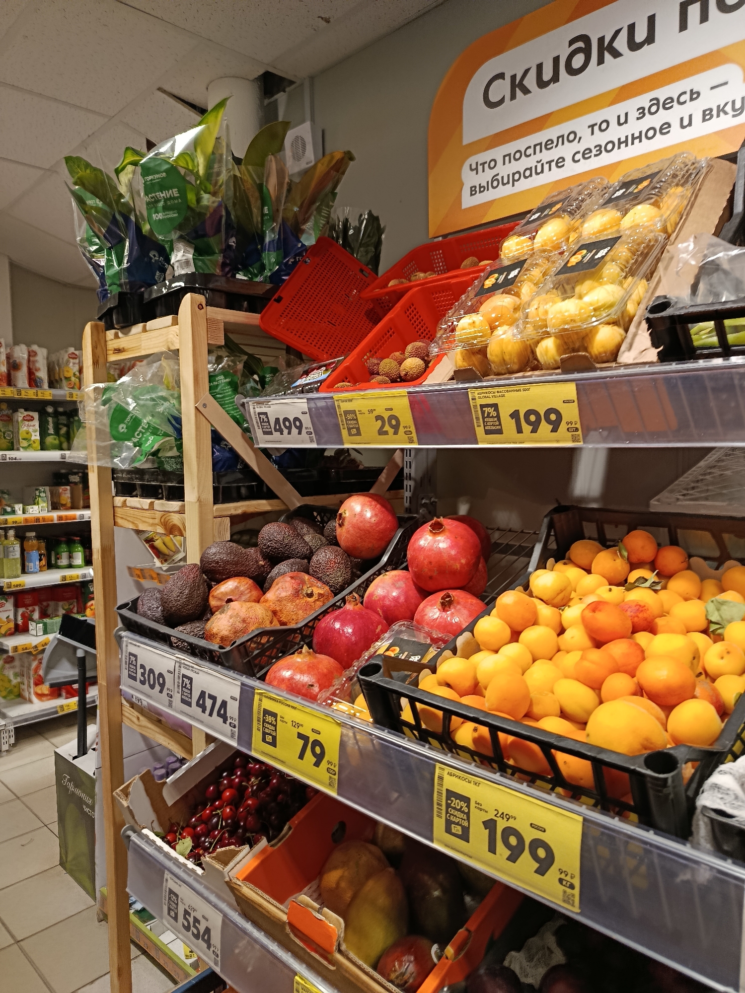A medium shot shows a grocery store aisle in Vereya, Russia, with shelves stocked with fruits and vegetables. The foreground displays an assortment of fruits, including pomegranates, avocados, and apricots, in black plastic crates. Yellow price tags with Russian text are attached to the shelves, indicating discounts and prices. A large orange and white sign with Russian text, reading "Скидки" (Discounts), is visible at the top of the image. The background reveals more shelves filled with packaged food items and leafy green plants. The lighting appears to be from overhead fluorescent lights. No people are visible in the frame.