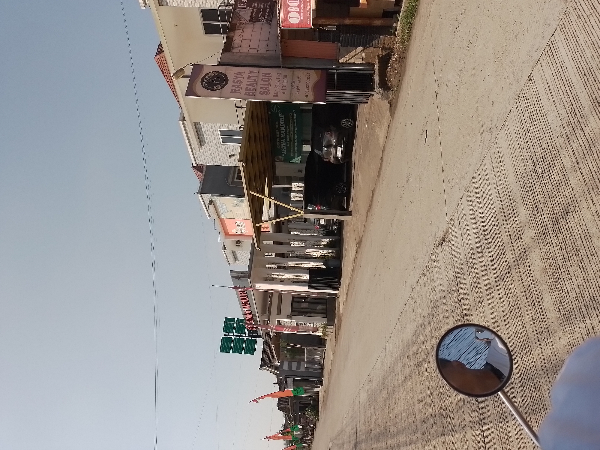 The image shows a street scene in Ngancar, Indonesia, on a clear day. On the right side of the street, there is a two-story building with "RAsya BEAUTY SALON" signage. The salon offers hair, body, face, and treatment services and is open from 08:00 to 18:00, with a phone number of 082230009574. Adjacent to the salon is a food stall with a red and white sign. Further down the street, a row of smaller buildings and houses line the left side, some adorned with flags. Several cars are parked in front of these establishments. The road is paved with concrete and shows some signs of wear. In the foreground, a motorcycle's rearview mirror reflects the rider and the sky. The overall scene depicts a typical commercial street in a residential area.