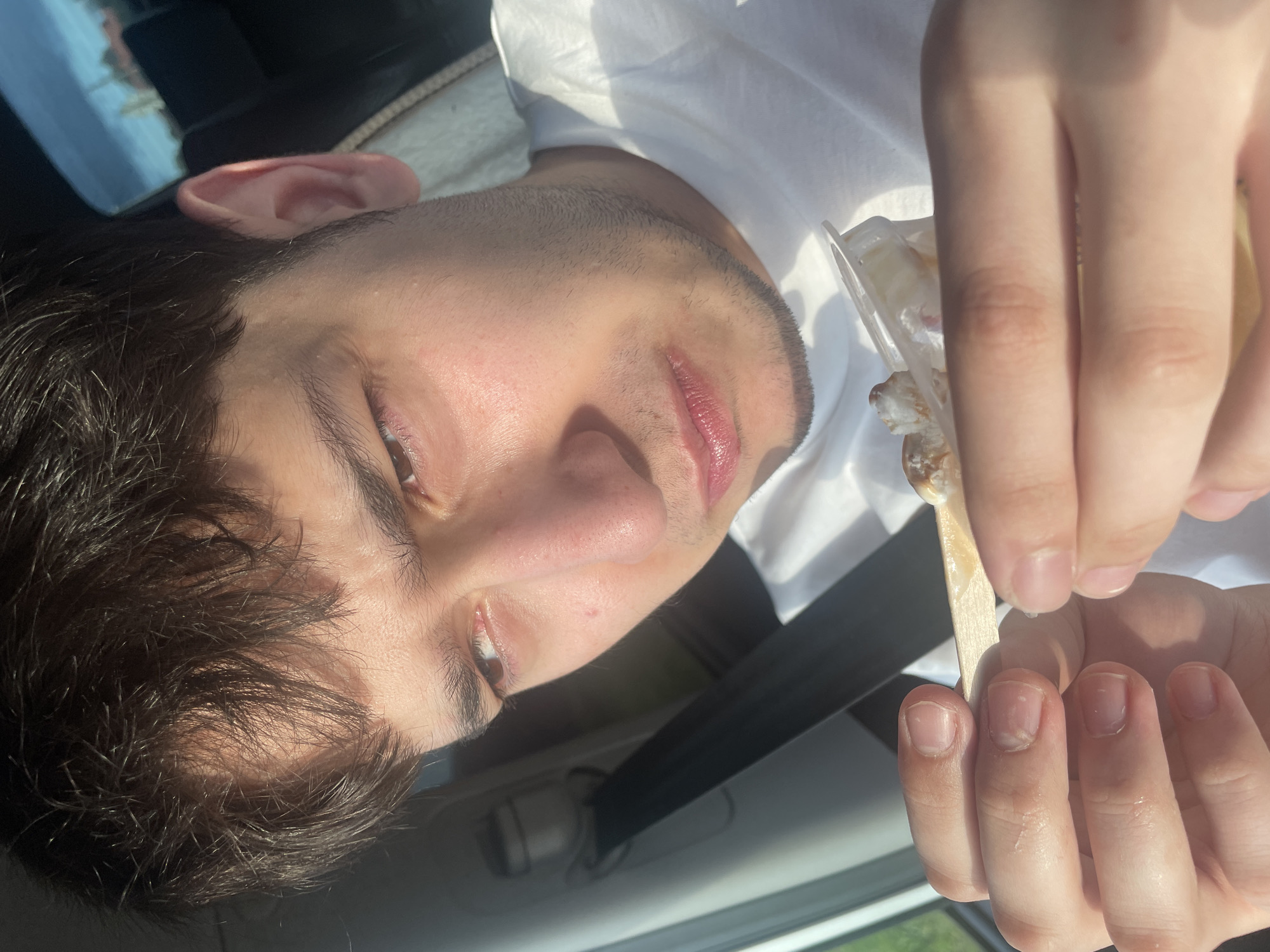 The image shows a close-up of a young person, possibly a male, inside a car. The person is looking towards the camera, with a portion of their face, including one eye, nose, and mouth, visible. They have dark hair and a light complexion. In the foreground, the person's hands are holding a small wooden stick with some white substance on it, possibly food or ice cream. One hand is touching the stick, while the other appears to be resting on the person's chin or neck area.
The setting appears to be daytime, with sunlight casting shadows, suggesting it might be a sunny day. The background shows a blurred view of the car's interior, including a seatbelt, and a reflection in a window that hints at an outdoor environment with some greenery. The overall mood seems casual, with the person possibly enjoying a snack. There is no visible text in the image. The provided location context "Гавриловка, Russia" suggests the image was taken in that region.