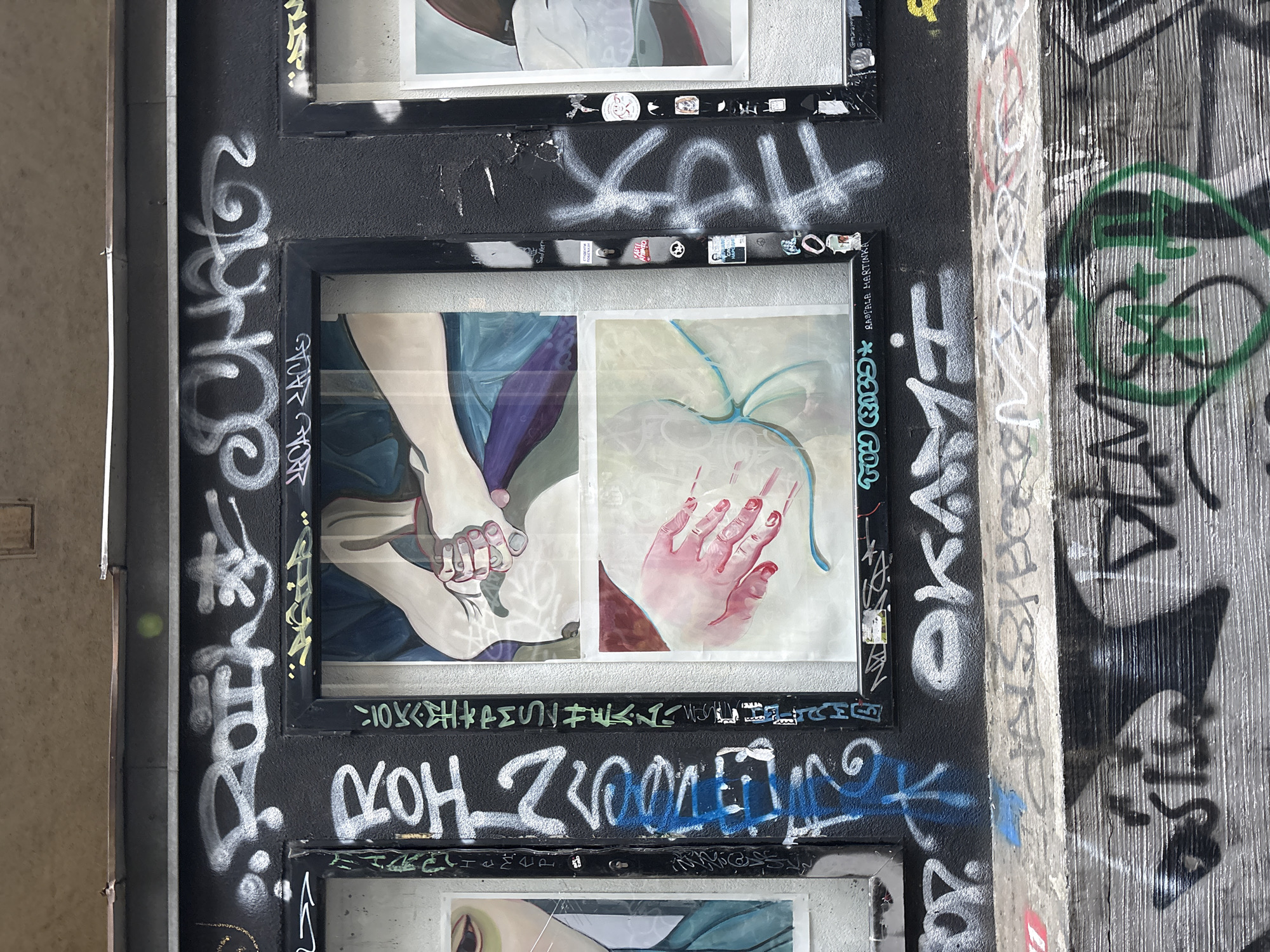 The image shows a section of an urban environment, likely a bridge support or wall in Belgrade, Serbia, heavily layered with graffiti. Several framed artworks are mounted on the black wall. The middle artwork depicts a pair of feet in a bluish color, and a hand in pink color. Graffiti is sprayed across the black wall and artwork frames. The frame on the right is tagged with "Basliela Martinka". The area to the right of the wall is covered with more elaborate, denser graffiti, featuring a green circle shape. The environment appears to be neglected.