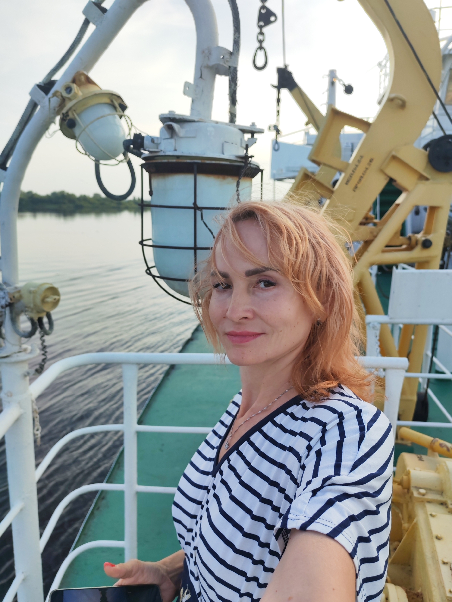 A middle-aged woman with light red hair and fair skin is taking a selfie on the deck of a ship. She is wearing a striped v-neck shirt. Behind her, the deck is lined with white railings, and a green painted surface is visible below. Various pieces of ship equipment, including a yellow crane and industrial lights, are present on the deck. The water of a river or canal is visible to the left, with a treeline on the distant shore. The sky is overcast. This scene is taking place on the Sheksna River, in or near the city of Sheksna, Russia.