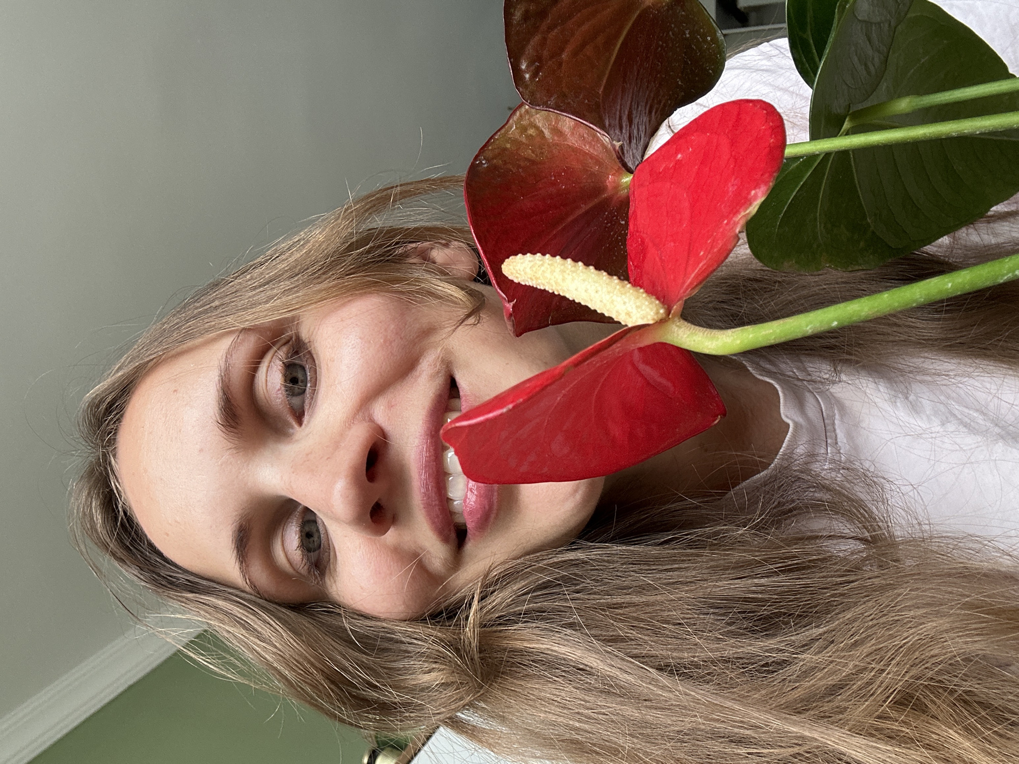 A fair-skinned woman with long, light brown hair is holding a flowering plant up to her face. The plant appears to be an anthurium with red and maroon spathes, as well as a green leaf. The woman is smiling. The background appears to be a gray wall. There is no visible indication that the photo was taken in Fethiye, Turkey.