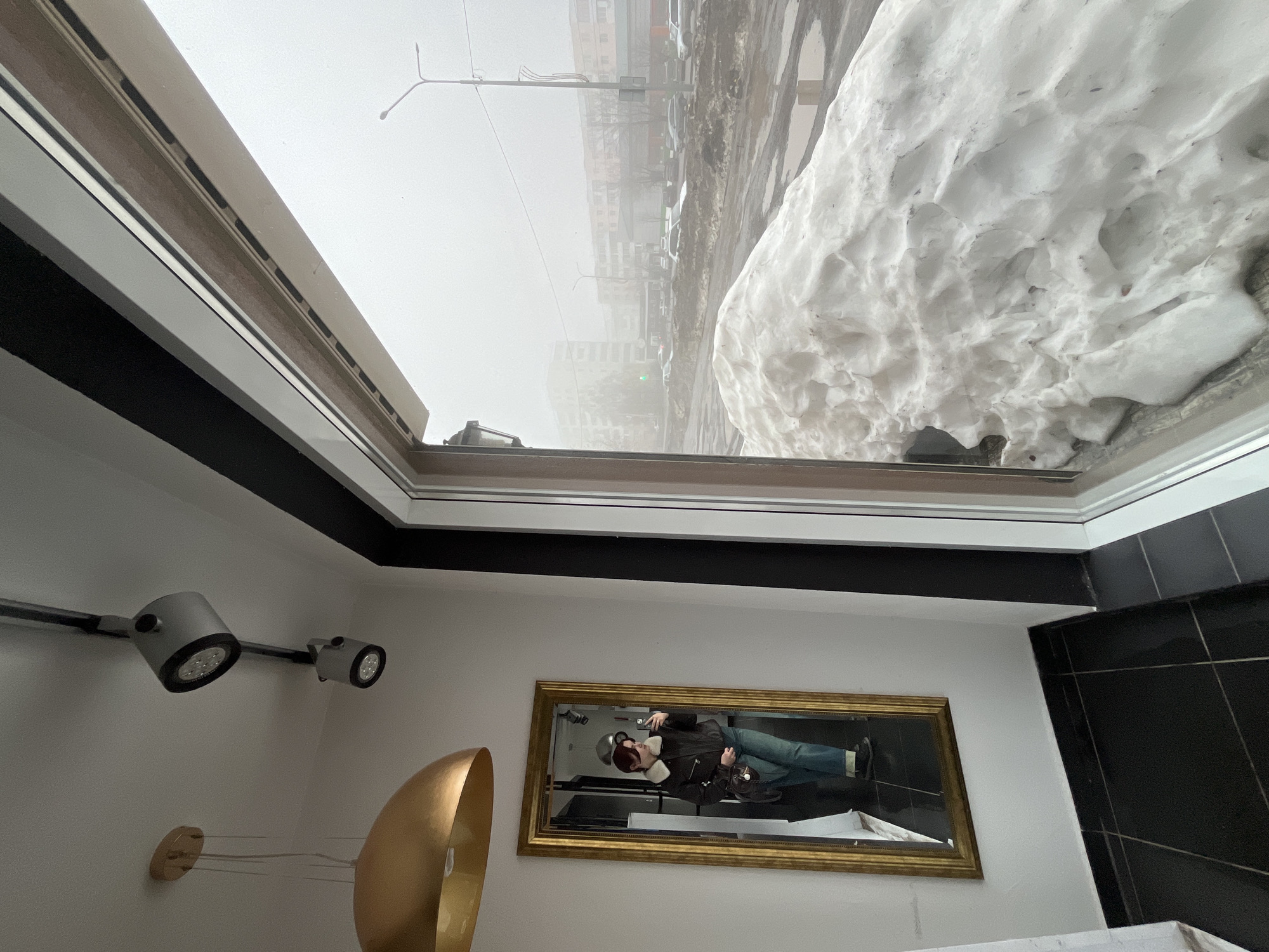 From an interior perspective, the scene shows a view through a window or skylight where heavy snowfall is visible outside. The location appears to be Neftekamsk, Russia, based on the urban landscape visible through the fog and snow. The cityscape includes buildings, a street with vehicles, and streetlights. Inside, there's a wall with track lighting and a gold-colored lamp. A mirror on the wall reflects a person holding a phone, apparently taking the photo of the scene. The black and white tiled wall further indicates an indoor setting.