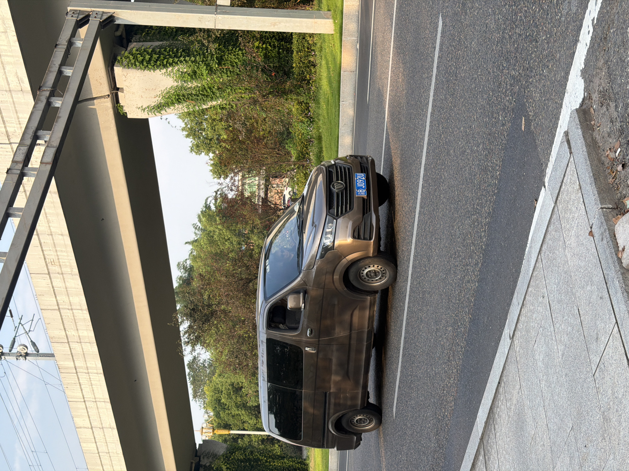A brown, four-door van is traveling on an asphalt road in Jiangjin, China. The van has tinted windows, silver rims, and a license plate reading "B-JO92U". The van is in the right lane, with a concrete sidewalk and pedestrian path to its left. The area is lined with green grass, trees, and foliage. Overhead, a concrete overpass supported by pillars stretches across the scene, with train tracks visible on top of it. The sky is clear and blue, with some overhead wires and utility poles. The lighting suggests it is late afternoon or early evening, as the sun casts long shadows. A person wearing sunglasses is visible in the driver's seat.