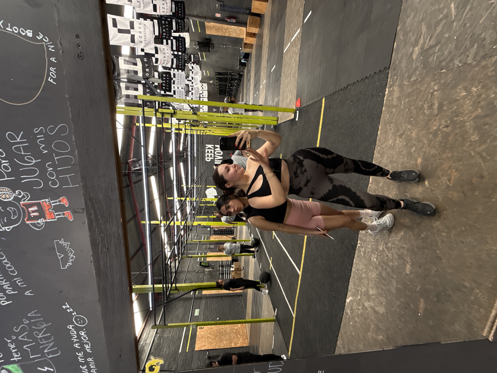 Two women pose for a selfie in an indoor gym or CrossFit box in Mexico City, Mexico. The woman in the foreground on the right takes the photo, holding a black smartphone. She wears a black sports bra and patterned dark gray leggings. Beside her, another woman looks towards the camera, dressed in a black top and light pink shorts. Both women appear to be smiling or content.
The setting features typical gym equipment, including metal racks and rigs that stretch into the background. The floor is a mix of black rubber mats with yellow demarcation lines and a lighter, distressed wooden or plywood section. In the background, other individuals are visible, some appearing to be exercising or preparing for workouts.
Notable details include banners or flags hanging from the ceiling, one of which clearly displays "HYROX" (visible correctly after rotating the image from the reflected "XORYH" in the OCR), "MEXICO", and "MONTEMORRO". The wall to the left is dark, resembling a chalkboard, covered in white chalk writing and drawings. Visible text on the wall includes "PARA JUGAR con mis HIJOS" (To play with my children), "tener MAS ENERGIA" (have MORE ENERGY), "QUE ME AYUDA A DORMIR MEJOR. :)" (That helps me sleep better. :)), and "FOR A NICE BOOTY". Bright overhead lights illuminate the space.