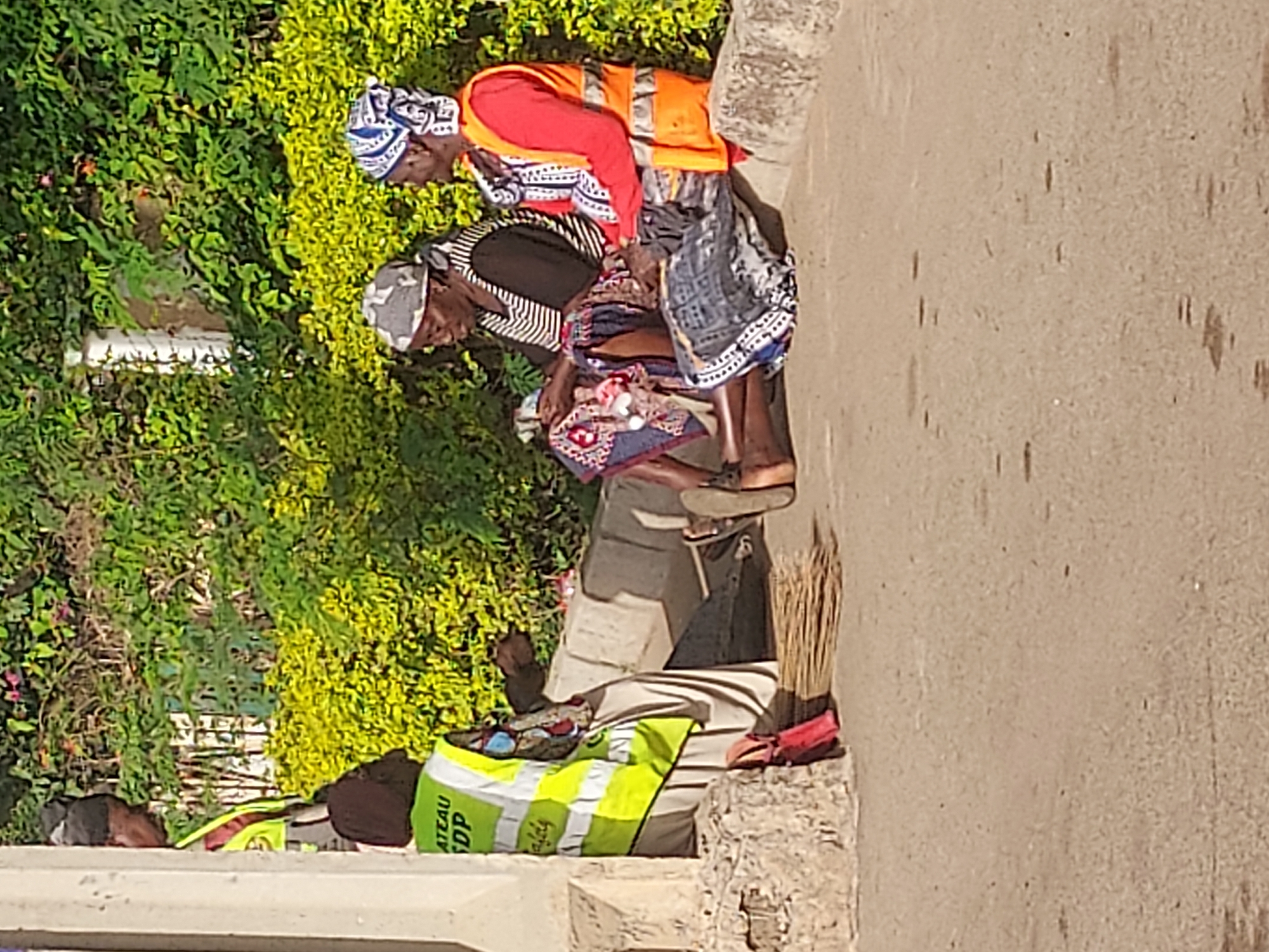 Two elderly women are positioned on a curb next to a road. One woman is wearing an orange vest over a red long-sleeved shirt and a patterned headscarf. The other woman is wearing a striped top, a patterned headscarf, and a patterned skirt. They appear to be examining or sorting through small items held in their hands. A bag with the markings "ATEAU SDP" sits nearby, along with a broom, both resting against the curb. Green foliage is visible in the background. The setting appears to be an urban roadside location in Gura Top, Nigeria.