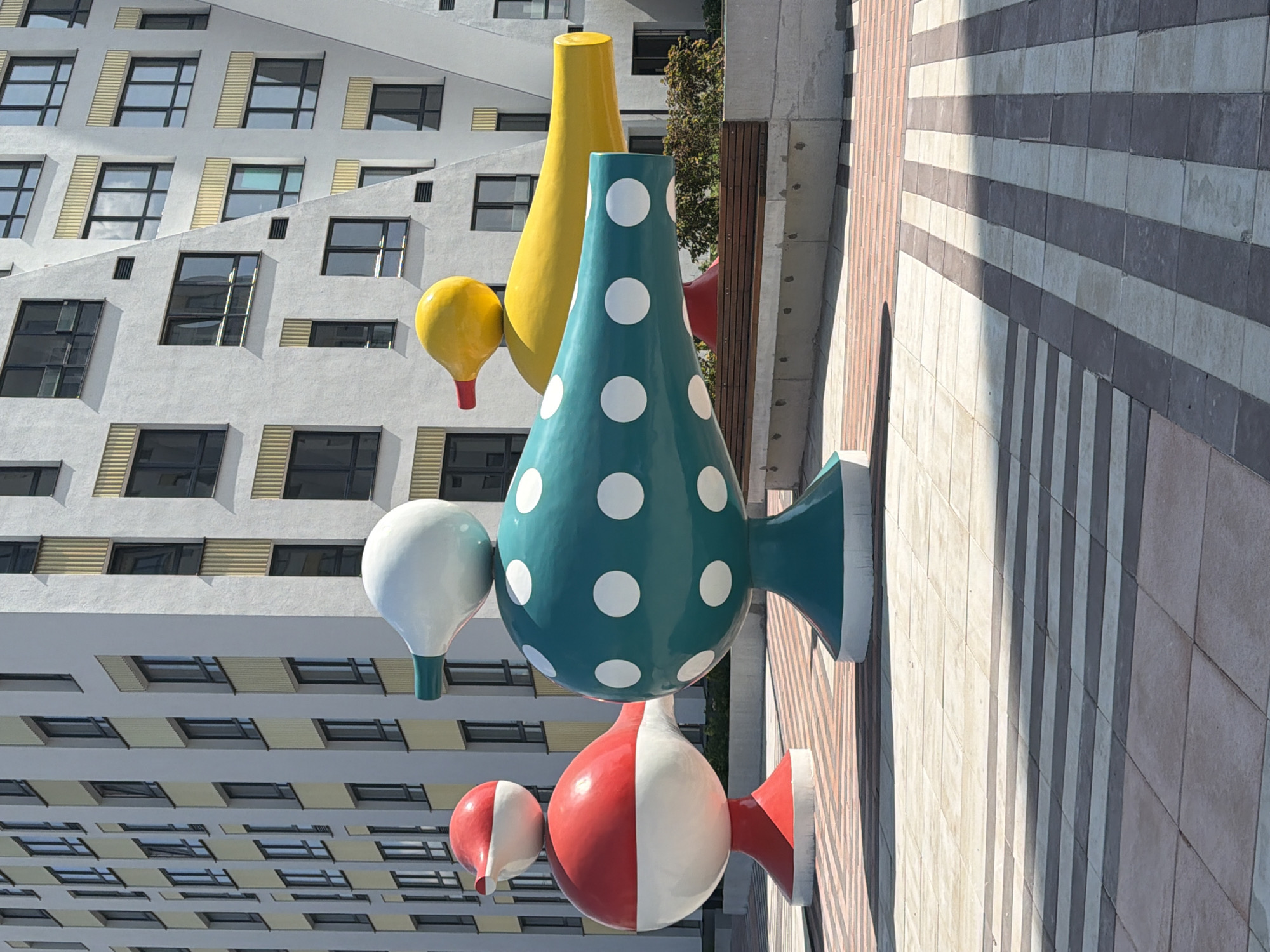 The image captures a public art installation featuring three whimsical, duck-like sculptures in Yekaterinburg, Russia. The sculptures are positioned in a row, with a red and white one on the left, a teal and white polka-dotted one in the center, and a yellow one on the right. They are made of shiny, smooth material and have rounded bodies with beak-like protrusions. The background is dominated by a modern, multi-story building with a light-colored facade and numerous windows. The ground in front of the sculptures is paved with geometric patterns of light and dark square tiles, with some striped tiling further back. A wooden bench is visible behind the sculptures. The lighting suggests it is daytime, with shadows cast by the sculptures and the building. There are no people visible in the image.