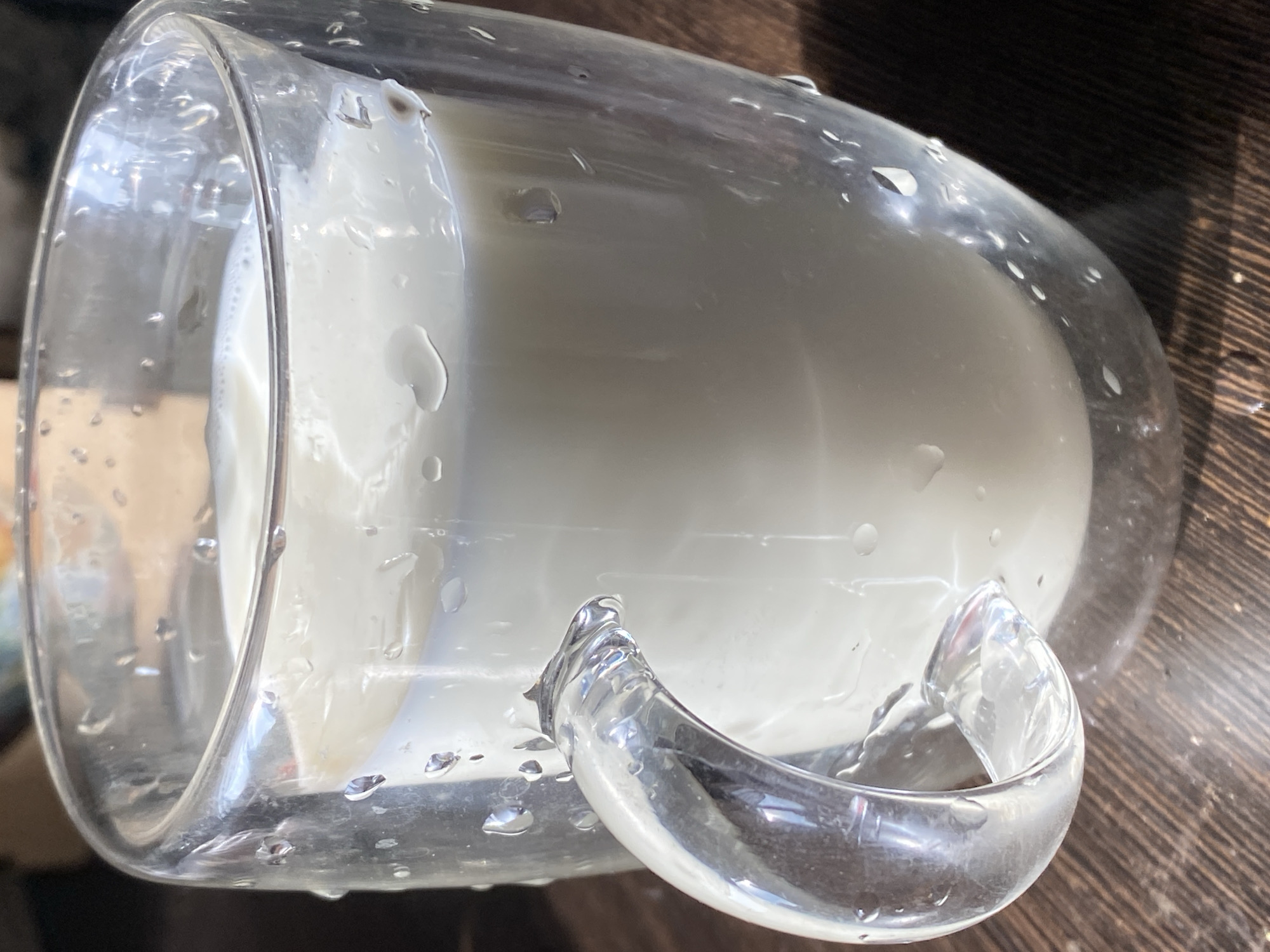 The image shows a close-up of a clear, double-walled glass mug filled with a white liquid, likely milk. The mug is placed on a wooden surface, which appears to be a table or countertop. The lighting suggests it might be daytime, with light reflecting off the water droplets that dot the exterior of the mug. These droplets, along with some condensation, indicate that the mug may have been recently filled or is holding a cold beverage. The mug has a clear glass handle attached to its side. The background is dark and out of focus, highlighting the mug and its contents as the primary subject. There is no visible text in the image. The scene is a simple, everyday object in a domestic or casual setting, possibly in Novokuznetsk, Russia, as per the location context.