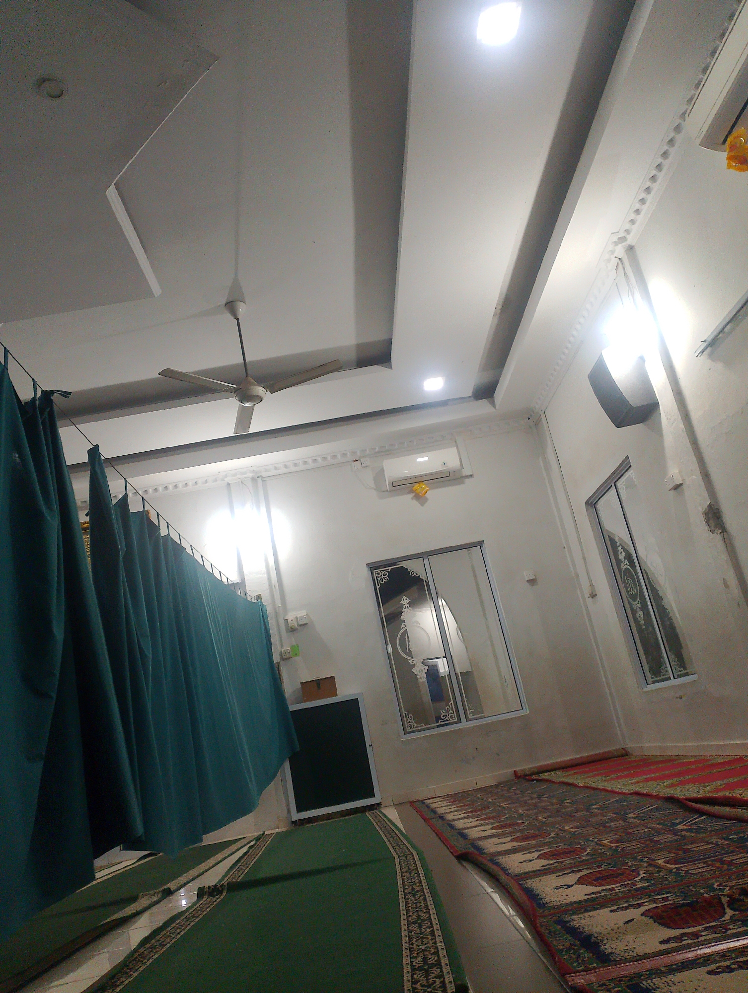 The image depicts the interior of a mosque, likely in Batam, Indonesia, featuring a prayer area with green carpets and prayer mats. A teal-colored curtain divides a section of the room, possibly for women. The ceiling has a modern design with recessed lighting and a ceiling fan. An air conditioning unit is mounted on the wall. Two large windows with decorative glass panels are visible. The overall environment appears clean and well-maintained, suggesting it is an active place of worship.