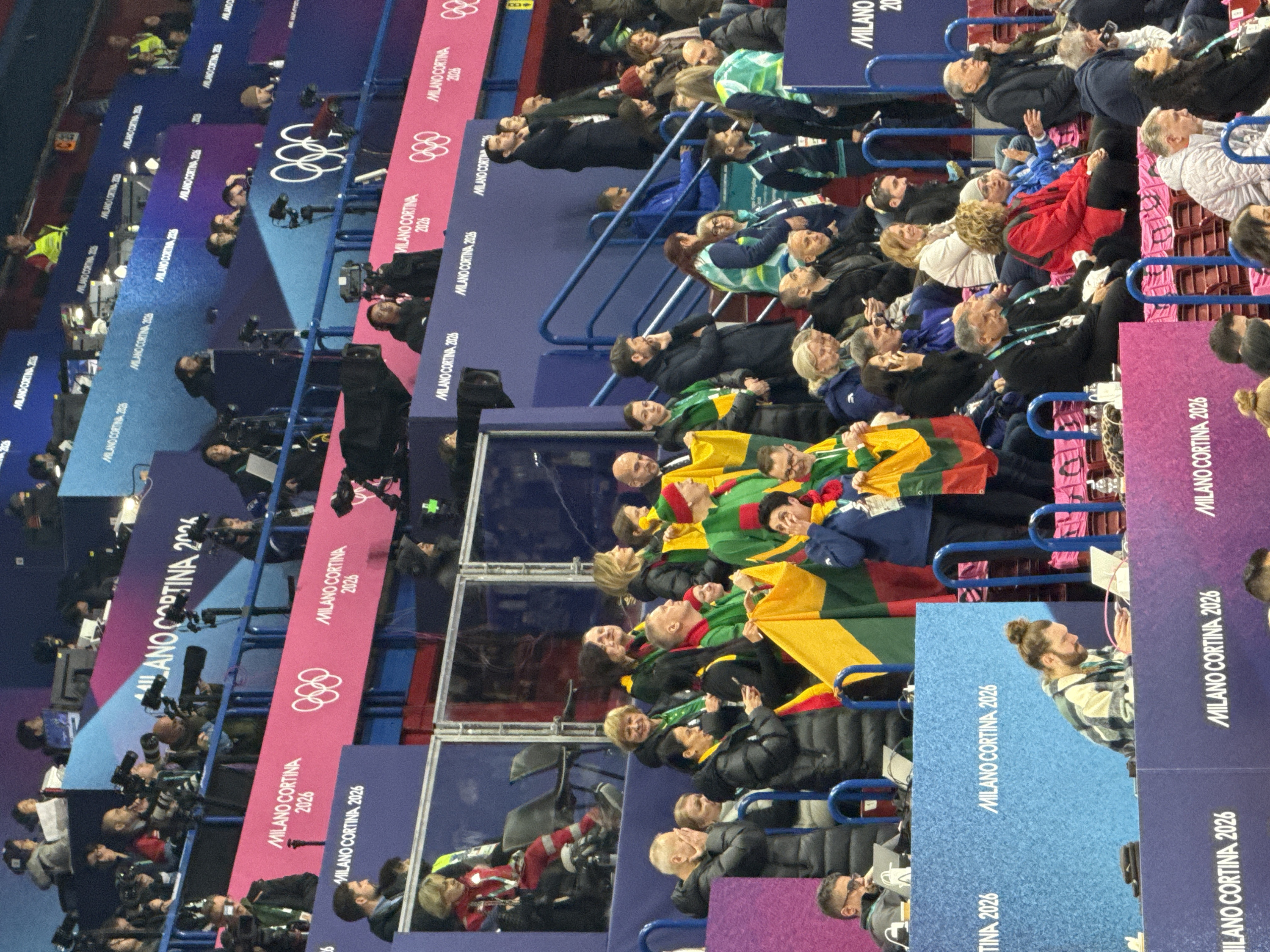 A busy scene shows a crowd of people at an event, possibly a sporting event or ceremony. Many people hold cameras, suggesting they may be journalists or photographers. A group holds up what appears to be a Lithuanian flag. Banners and signs with the words "Milano Cortina 2026" and the Olympic rings are visible, which indicates this event is likely related to the 2026 Winter Olympics in Milan and Cortina d'Ampezzo. The crowd is seated on rows of seats in a stadium or arena.
