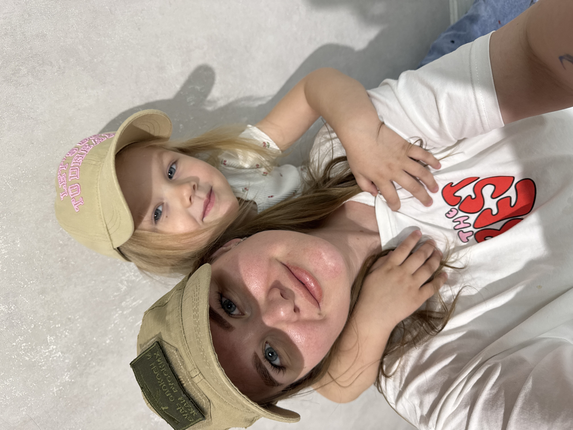 Two individuals, a woman and a young child, lie side-by-side. Both are wearing light-colored baseball caps. The child's cap has pink lettering. The woman's cap has a green patch. Both are wearing white shirts. The child is partly on top of the woman, with her arm around her. They appear to be indoors. The image was taken in a residential setting, likely within the city of Gornyi (Dvurechenskoe S/P), Russia.