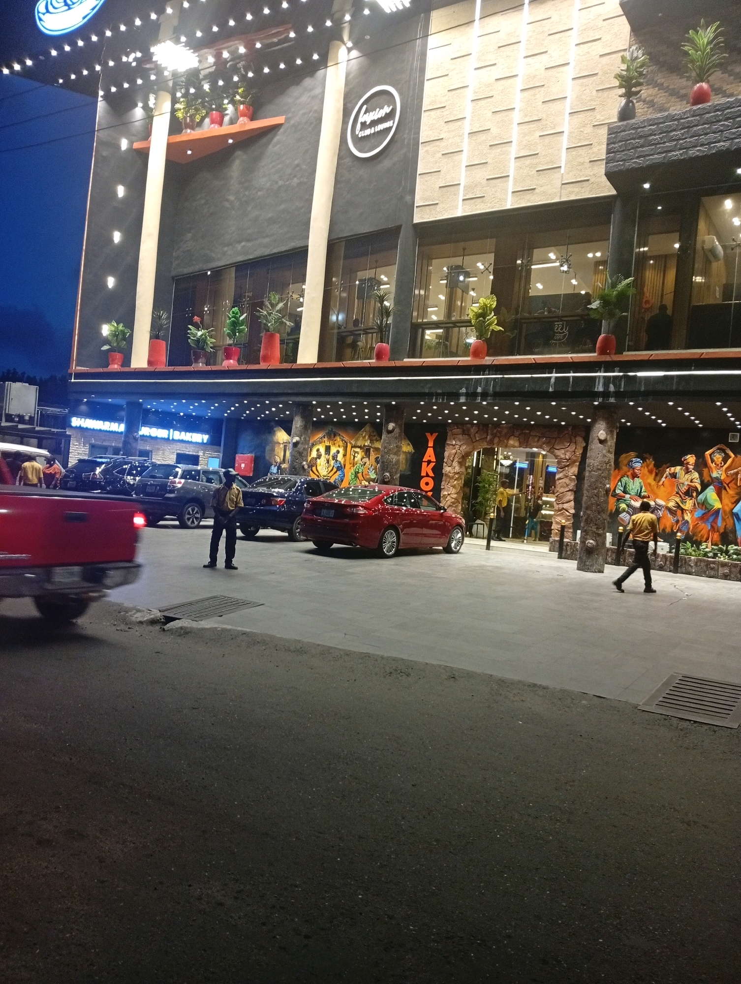 The image depicts the exterior of a multi-story building at night, likely a restaurant or entertainment venue, located in Ikeja, Nigeria. The building features a modern facade with illuminated signage and decorative lighting. Below the main structure, there's a covered walkway with parked cars and a large mural on the wall.
In the foreground, a red pickup truck is partially visible on the left, with motion blur indicating it might be moving. Several other cars are parked in front of the building.
There are at least three individuals visible. One man, wearing a yellow shirt and dark pants, stands in the middle of the paved area, looking towards the building. Another person, dressed similarly, walks away from the building on the right side of the image. A third individual, also in a yellow shirt, is seen through the glass doors of the building.
The environment suggests an urban setting at dusk or nighttime, with artificial lighting illuminating the scene. The building appears to be a commercial establishment.