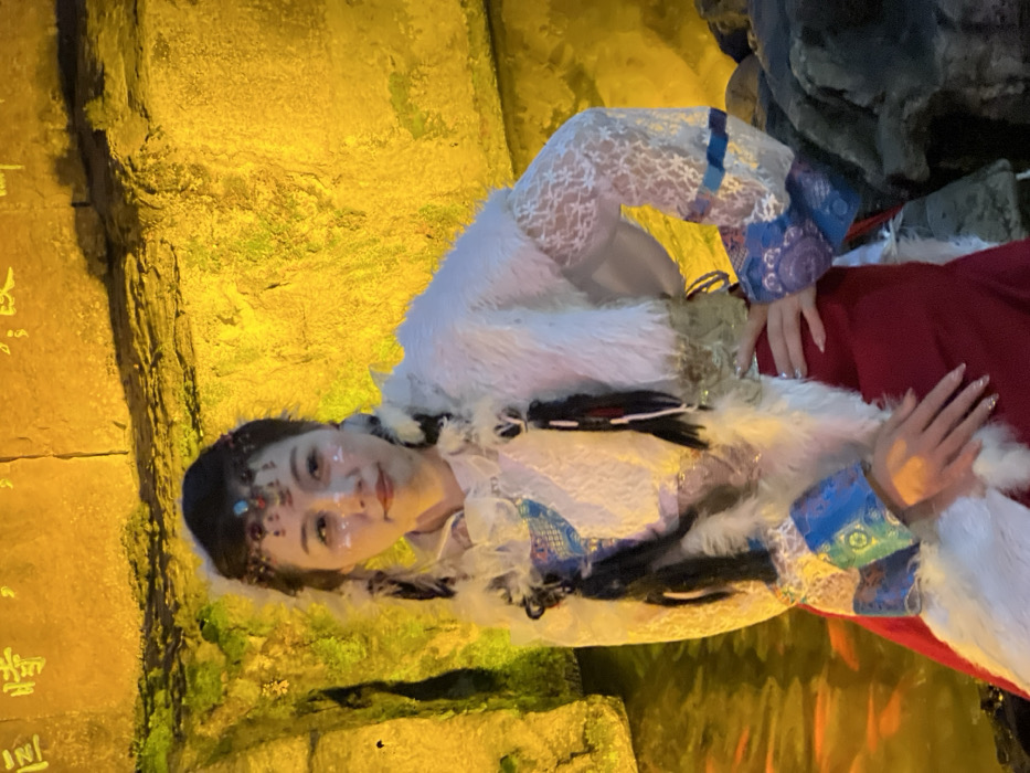 A female individual, appearing to be of East Asian descent, stands in a staged indoor environment located within Yuzhong District, China. She wears a traditional or theatrical costume featuring a white, fur-like sleeveless vest over a long-sleeved, translucent white lace garment with patterned blue and gold cuffs. A red lower garment or skirt is also visible. Her dark hair is styled into two long braids, each adorned with white feathers and black tassels. A beaded headpiece, featuring a central blue-green stone and small white feathers, is worn on her forehead. Her face has light makeup, including small white, reflective accents on her cheeks and forehead. Her fingernails are long and appear to have a light, possibly glittery, polish. Her left hand rests over her right forearm, and her right hand is positioned on her hip. She faces slightly right of the viewer, looking towards the camera with a neutral expression.
The background consists of a textured rock wall, heavily illuminated with a warm yellow light. Indiscernible Chinese characters are visible on the upper section of this wall. To the subject's left, a body of still water reflects the yellow light and contains several visible orange fish, consistent with koi. To her right, darker, uneven rock formations are present. The scene is dominated by a strong, artificial yellow illumination.