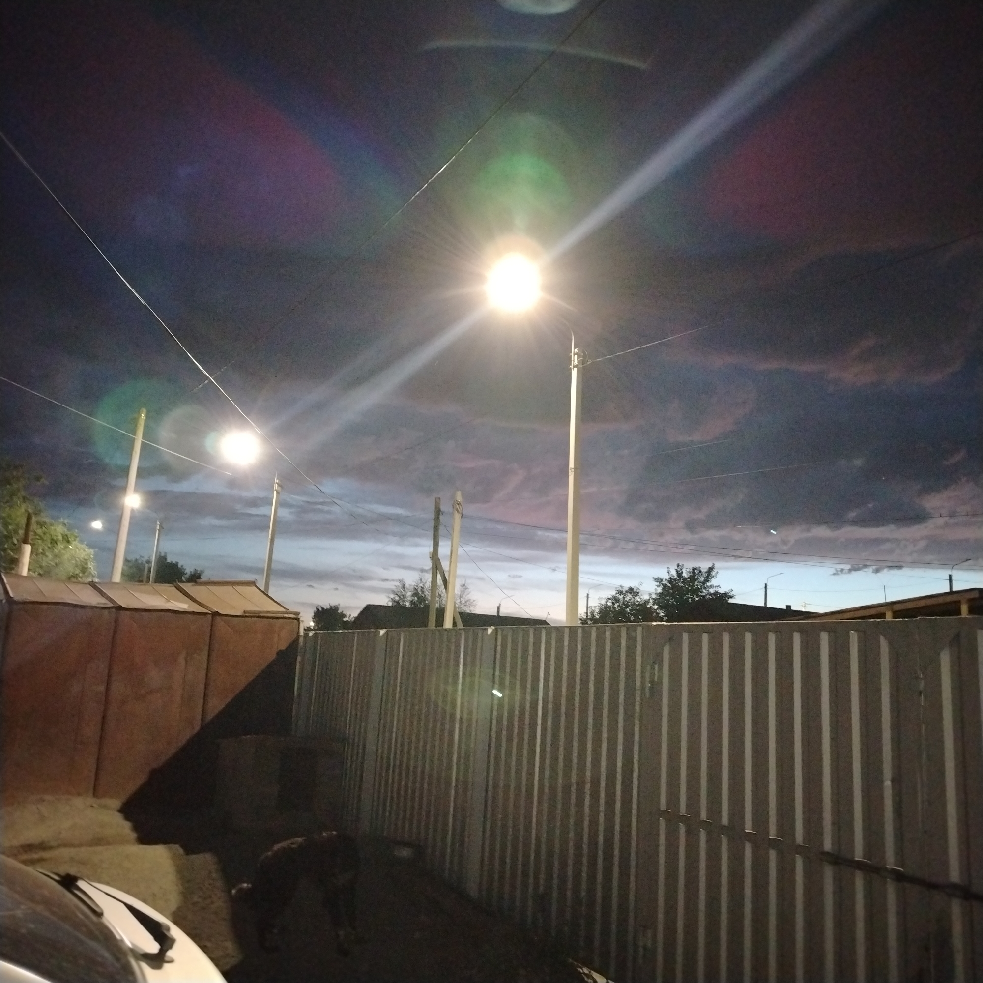 This media file shows an outdoor scene at dusk in Ekibastuz, Kazakhstan. The dominant feature is a street lamp illuminating the twilight sky, which is filled with dark, dramatic clouds tinged with pink and purple hues. Two tall lampposts are visible, one with a bright, blown-out light source at its peak and another with a smaller, less intense light. Wires crisscross the sky, connecting to the lampposts and disappearing into the frame.
In the foreground, a corrugated metal fence runs horizontally across the image. To the left of the fence, a large, brown, likely industrial or storage structure is partially visible, casting a long shadow. Behind the fence, more trees and the roofs of buildings can be seen in the distance, suggesting a semi-urban or industrial area. The overall mood is somewhat somber, with the dark clouds and artificial lighting creating a dramatic and slightly eerie atmosphere. The time of day is clearly evening, as indicated by the street lights and the twilight sky. No text is visible in the image.