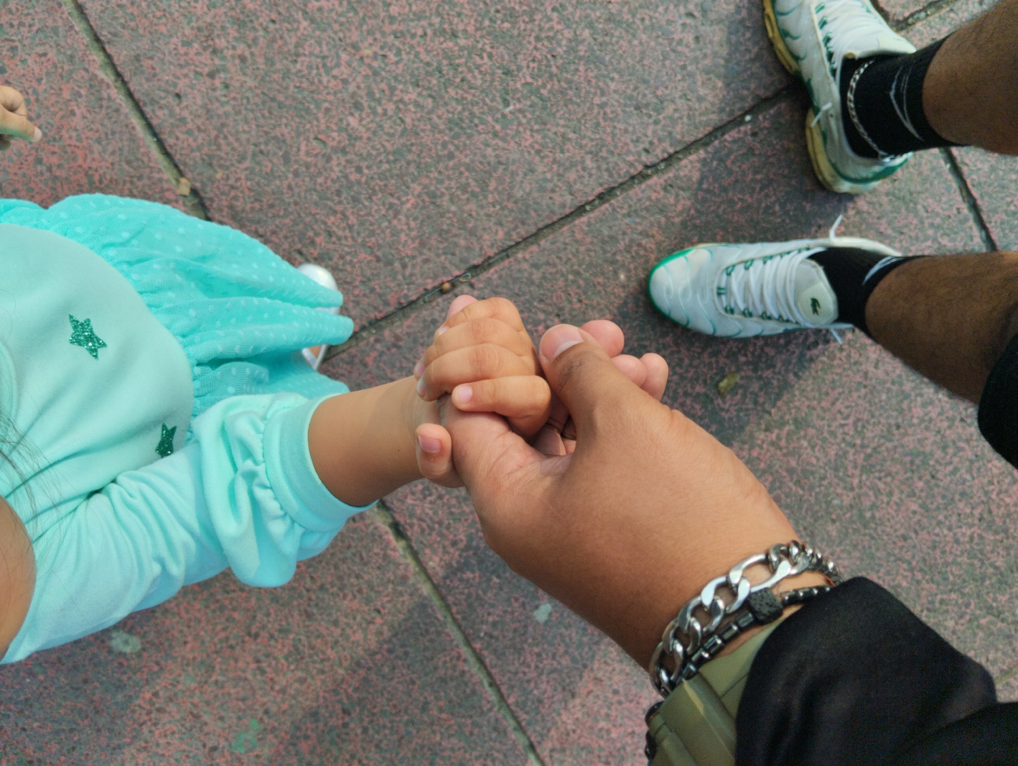 An overhead view captures an adult holding the hand of a child on a textured, reddish-brown and grey paved surface, likely a sidewalk or public area in Caracas, Venezuela. The child, partially visible on the left, wears a light blue long-sleeved top with a sparkly green star appliqué and a light blue polka-dotted skirt or dress. The adult's arm, dressed in a black sleeve, features a prominent silver chain-link bracelet, a dark beaded bracelet, and a partially visible olive green watch. Below, the adult's legs are seen wearing black socks and white and green sneakers, one of which displays a small green crocodile logo on its tongue. The bright lighting suggests it is daytime. The gesture of holding hands conveys a moment of connection, guidance, or care between the two individuals.