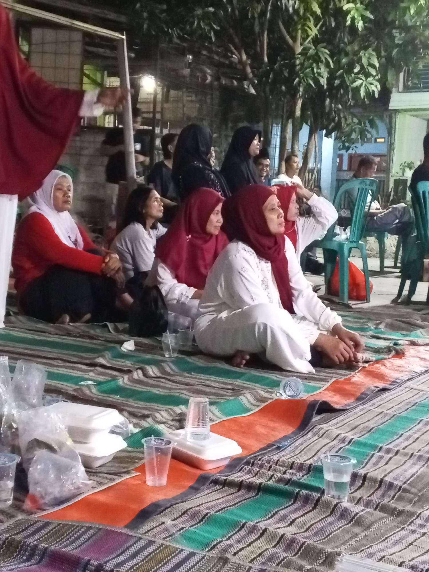 In Ciruas, Indonesia, at night, a group of people, predominantly women in hijabs, are gathered outdoors on a patterned mat. They are seated on the mat, some with their legs crossed, suggesting a communal gathering. There are several white styrofoam food containers and plastic cups scattered on the mat, indicating that a meal or refreshments have been served or are being consumed. Behind the seated individuals, other people are visible, some standing and some seated on plastic chairs. Trees and some structures, possibly buildings, form the background. The lighting appears to be artificial, possibly from overhead lamps or building lights. The overall scene depicts a casual evening gathering in an outdoor setting.