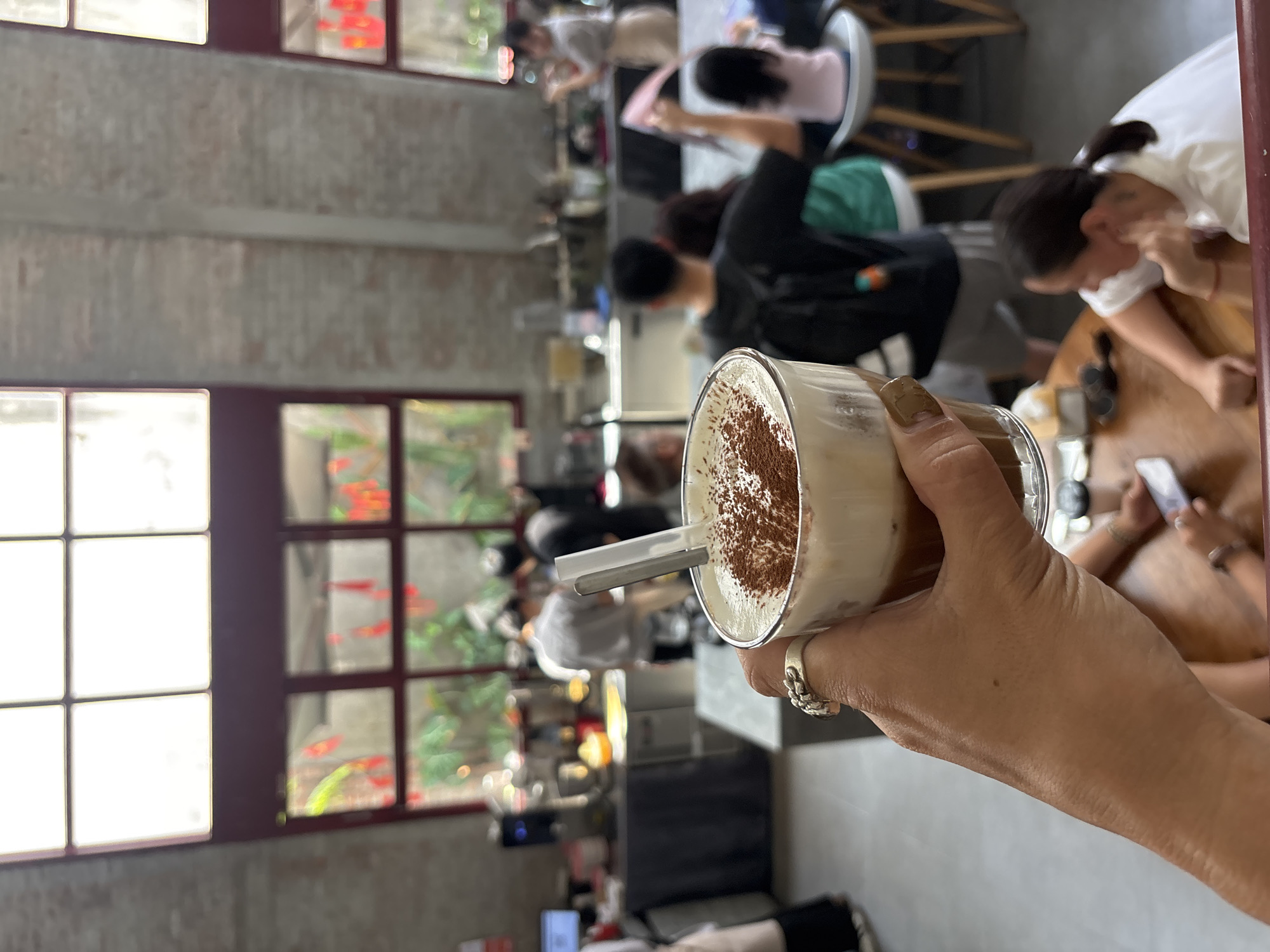 A close-up shot shows a hand holding a glass of coffee topped with whipped cream and dusted with cocoa powder. The background is blurred, revealing a cafe interior with patrons and baristas. Large windows with red frames offer a glimpse of greenery outside. The setting appears to be a modern cafe in Đà Nẵng, Vietnam, with exposed brick walls and industrial-style lighting. The hand has a ring on the index finger and a nail with light brown polish. The overall atmosphere suggests a casual and lively environment.