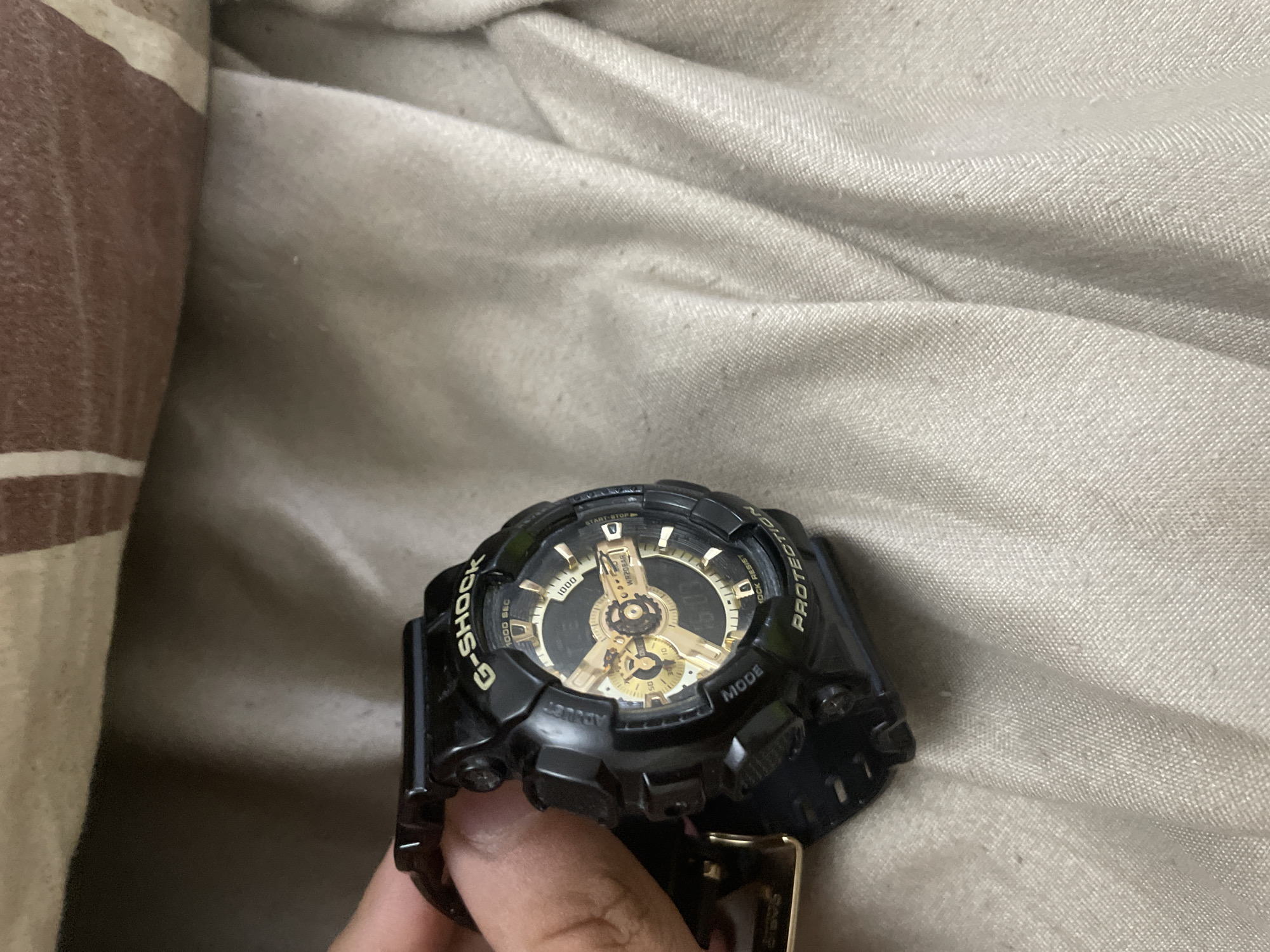 The image primarily features a black G-Shock watch with metallic gold accents, held by a person's fingers. The watch has a robust, sporty design, typical of the G-Shock brand, with a complex dial displaying various sub-dials and hands in gold. The watch is positioned against a background of fabric, suggesting an indoor setting.
Visible text on the watch includes "G-SHOCK" prominently on the left side of the bezel, and "PROTECTION" and "SHOCK RESIST" on the right. Other labels seen are "ADJUST", "MODE", and "START-STOP". The watch face also displays "WR20BAR" (indicating 20 Bar water resistance) and numerical markers such as "1000 SEC", "1000", "50", "10", and "000".
The background consists of two distinct fabric textures. On the right, a light, neutral-colored (possibly off-white or light gray) fabric with soft folds and a subtle weave is visible. On the left, a section of striped fabric, featuring reddish-brown and off-white bands, provides a contrast. The lighting appears somewhat dim, suggesting an indoor environment. A human hand, holding the watch, is partially visible at the bottom of the frame. No specific activity other than holding the watch is depicted, and no emotions are discernible. The location context provided is Ihtiman, Bulgaria, though no visual elements within the image directly confirm this geographical setting.