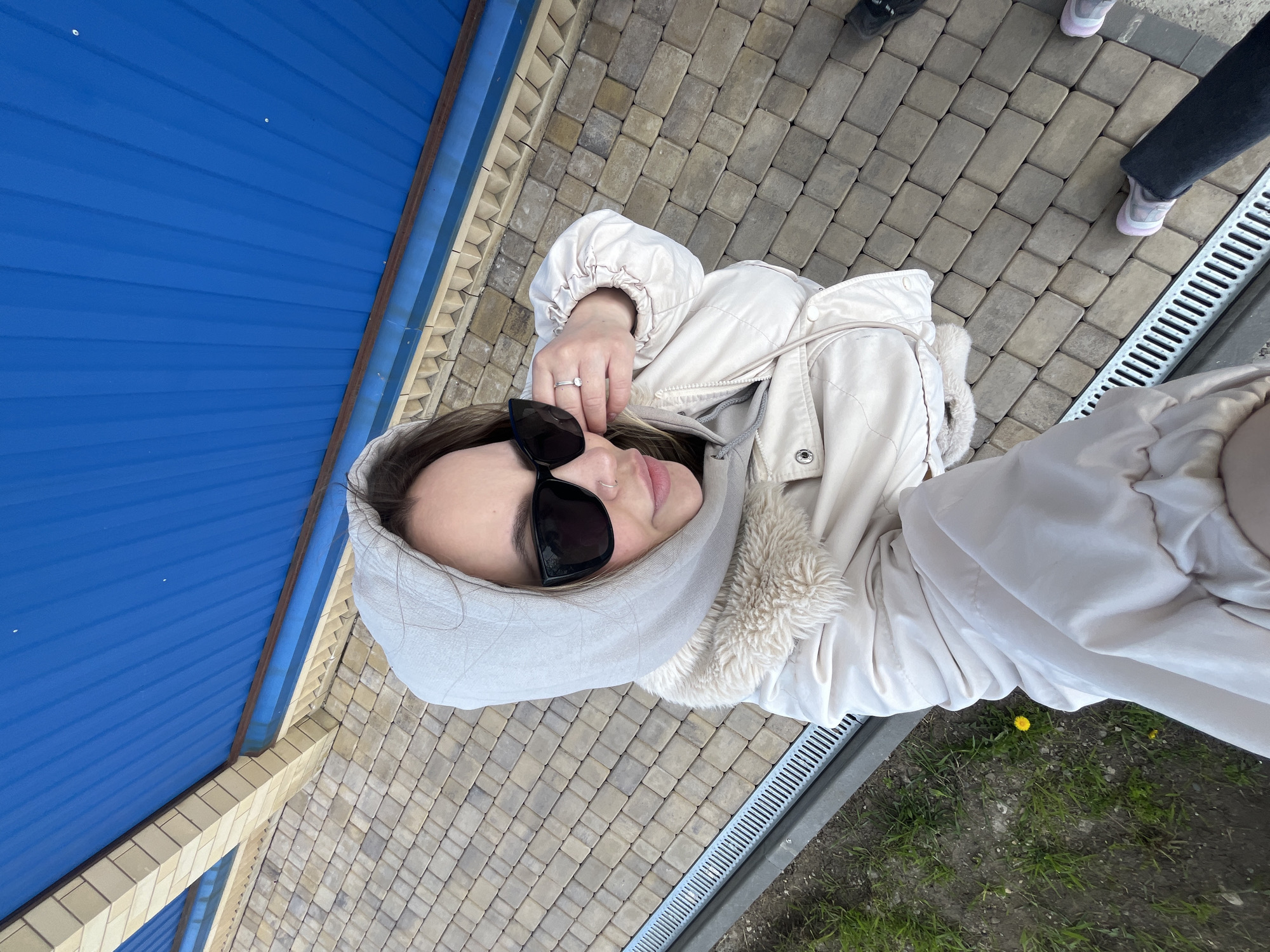 A woman wearing sunglasses and a hooded jacket is taking a selfie. She is standing on a paved surface, possibly a sidewalk or courtyard, next to a blue building. Two other people's feet are visible in the upper right corner of the frame. There is a metal grate and a patch of grass with a dandelion visible on the right side. The location is Novopokrovskaya, Russia.