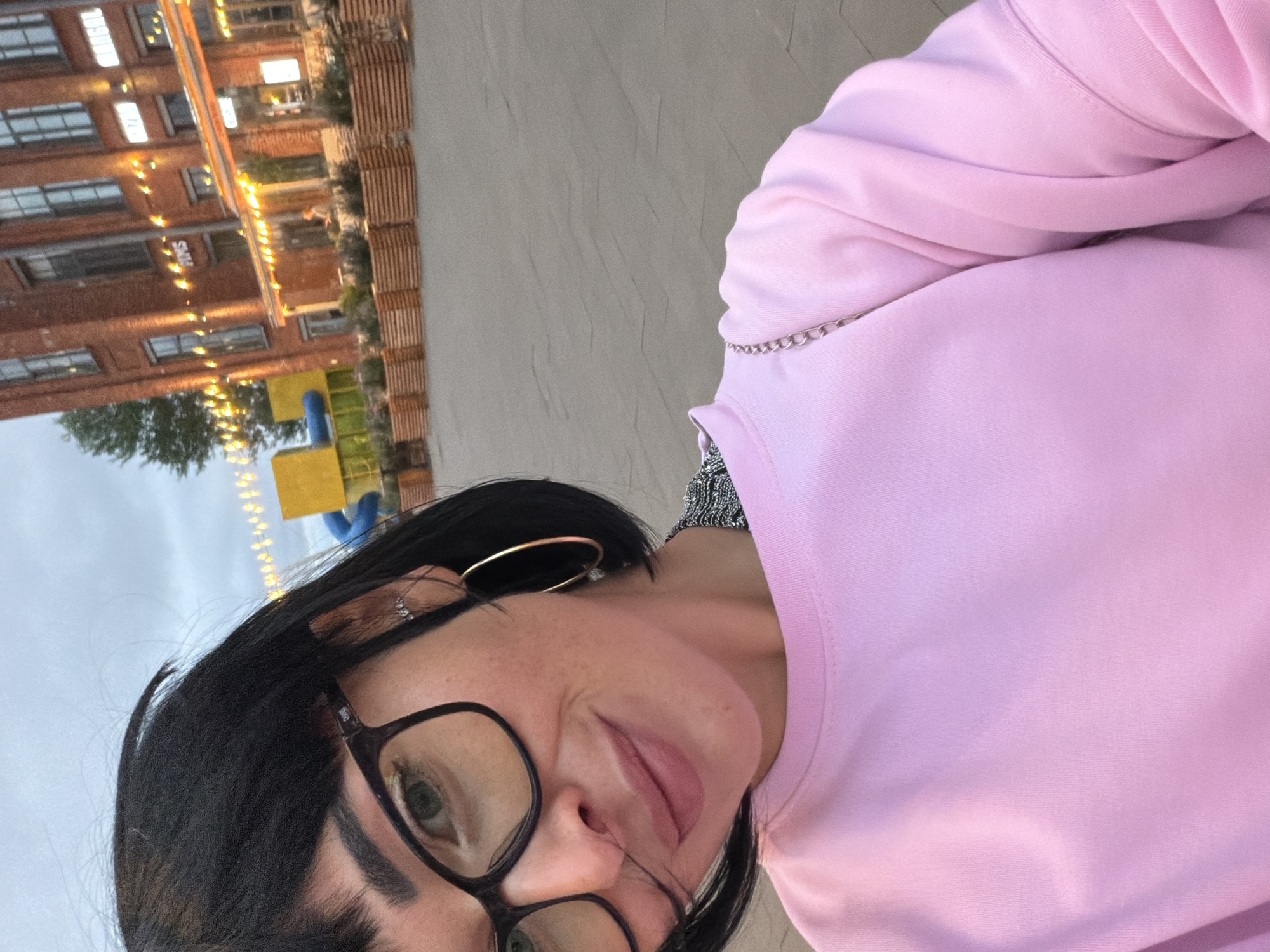 A woman with short black hair and bangs, wearing black-rimmed glasses, a pink sweatshirt, and large hoop earrings, smiles at the camera. She is standing outdoors in what appears to be a public square or pedestrian zone. In the background, a red brick building with illuminated windows and string lights is visible. Wooden planters filled with greenery line the front of the building. To the left, a bright yellow structure with a blue slide and green accents suggests a playground or public art installation. The ground is paved with grey cobblestones. The lighting indicates it is late afternoon or early evening. The scene is likely from a modern, revitalized urban area. Given the architectural style and the presence of a playground, this could be an outdoor area within the Sevkabel Port complex on Vasilyevsky Island in Saint Petersburg, Russia, which is known for its industrial-chic aesthetic, public art, and recreational spaces.