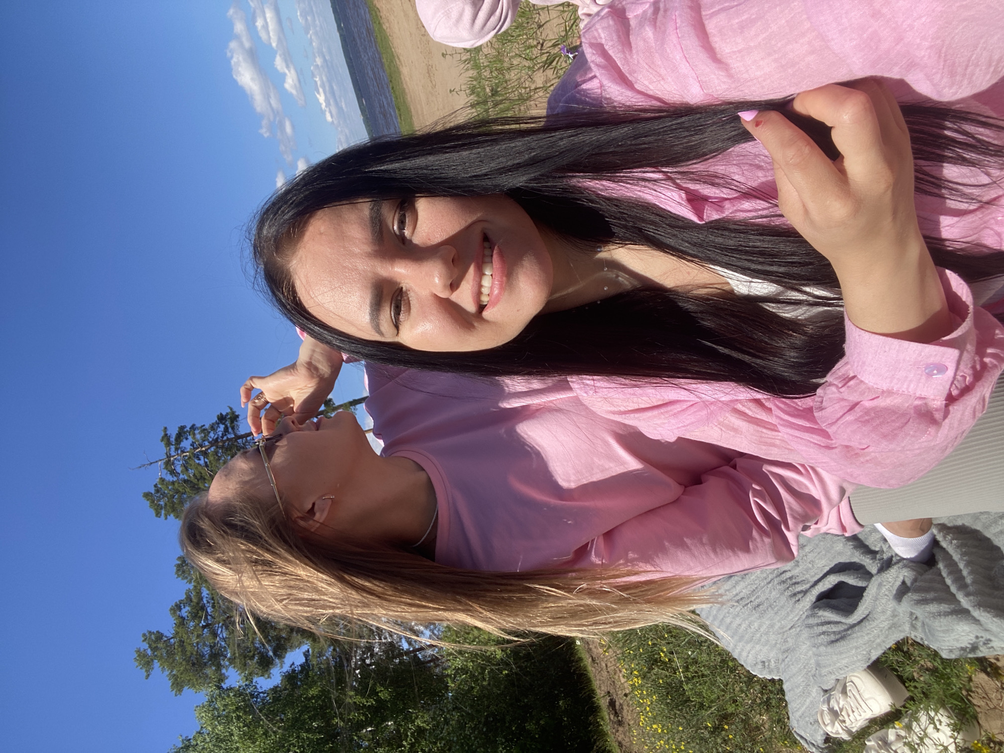 The image shows two young women in pink tops posing for a selfie outdoors on a sunny day. The woman in the foreground has long black hair and is smiling broadly, looking directly at the camera. The woman behind her has long blonde hair and is looking to her side, adjusting her sunglasses. They are sitting on a gray blanket in a grassy area with yellow wildflowers. In the background, there is a body of water, a sandy shore, and trees under a bright blue sky with scattered clouds. The location is likely a park or beach area near Chaikovsky, Russia.