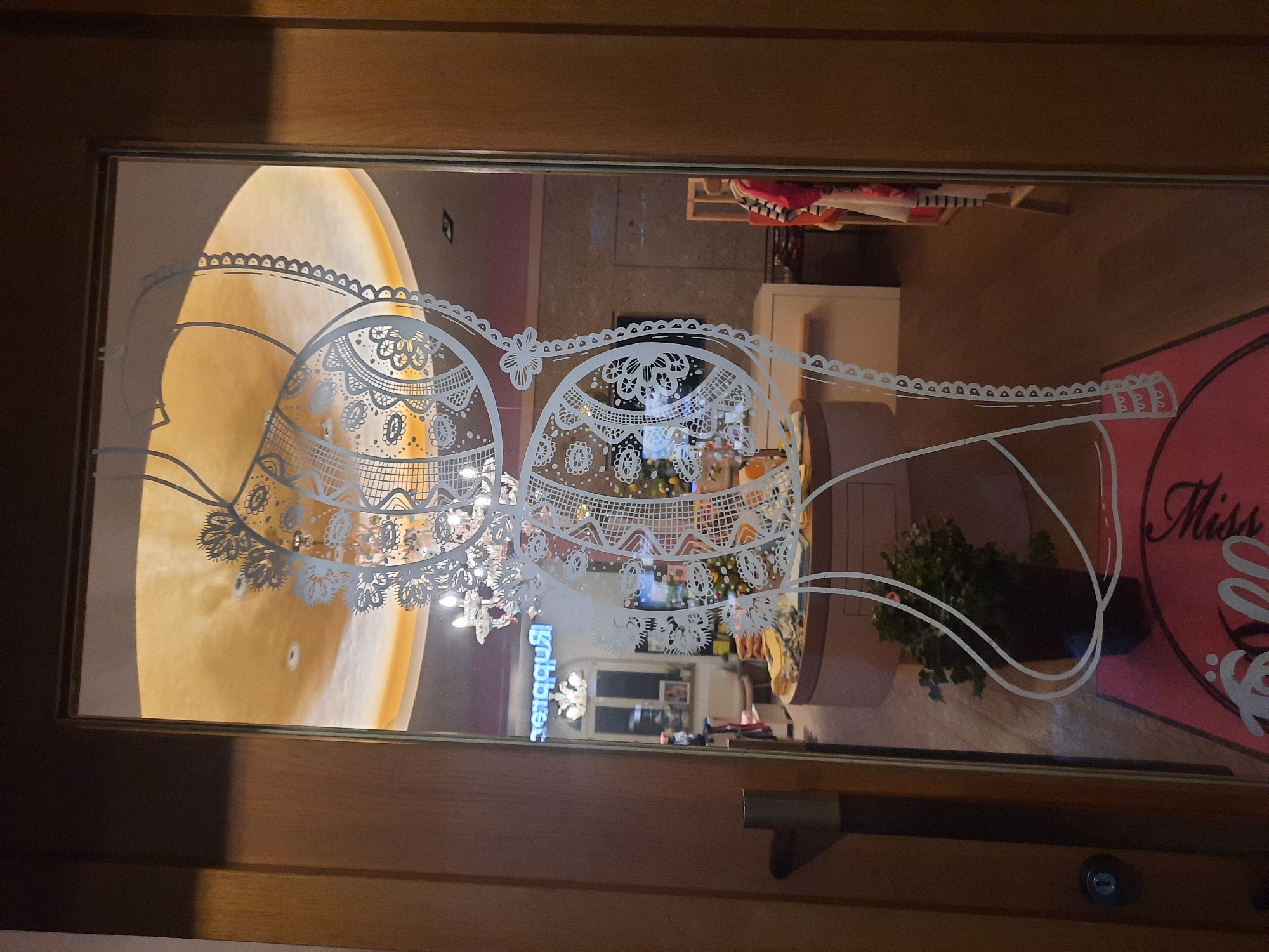The image shows the reflection in a glass door, which is decorated with a white lace-like pattern. The interior of a store is visible through the glass. The store appears to be a clothing boutique. There is a display counter in the center of the store, with some merchandise on it. A potted plant is on the floor to the left of the counter. Clothing racks are visible in the background. A neon sign that reads "Rupprecht" is reflected in the glass. The door has a wooden frame and a silver handle. On the floor in front of the door is a pink mat with white text.
This image was taken in Rosenheim, Germany, at the clothing store Rupprecht.
