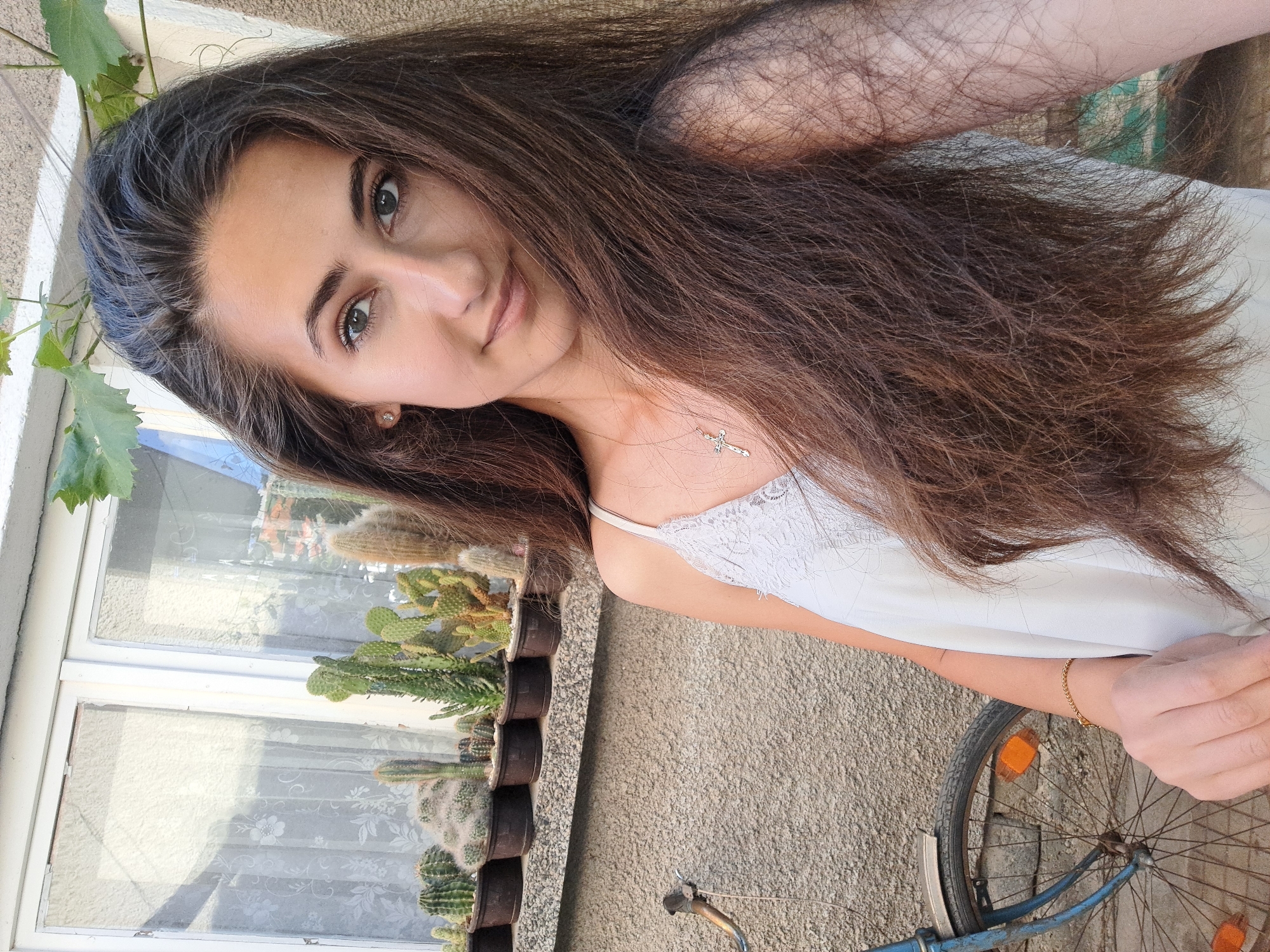 A young woman with long, dark brown hair is captured in a selfie outdoors. She is wearing a light-colored, thin-strapped top with lace detailing. Around her neck, she wears a delicate silver necklace with a cross pendant. Her left arm, adorned with a thin gold bracelet, is partially visible in the foreground.
Behind her, a row of potted cacti sits on a ledge beneath a window with white frames and sheer, patterned curtains. The wall behind the cacti is a textured, beige concrete. To the left of the frame, the front wheel and handlebars of a blue bicycle are visible, leaning against the wall. The bicycle has orange reflectors on its wheel.
The overall setting suggests an outdoor space, possibly a yard or patio, with hints of a residential building in the background. The lighting indicates it is daytime, with sunlight casting shadows and highlighting details. The location is identified as Chepintsi, Bulgaria.