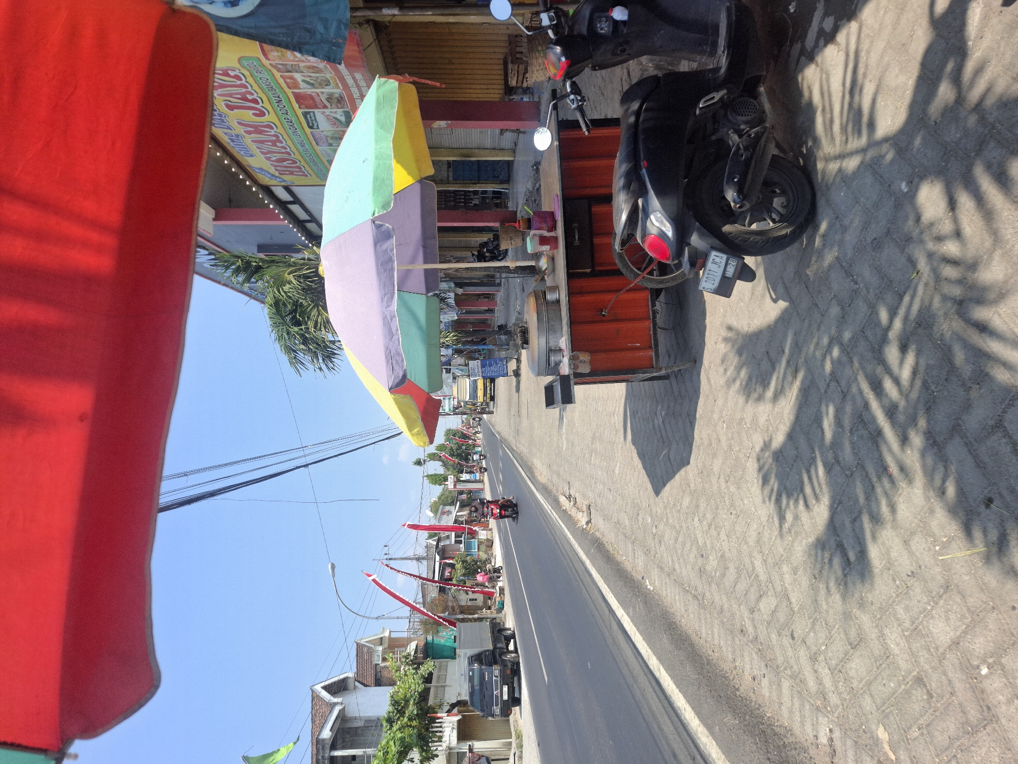The image depicts a street scene in Karanggeneng, Indonesia, on a bright, sunny day. A paved road runs horizontally across the frame, with a sidewalk on the right. Several red and white flags are visible along the roadside, indicating a potential local celebration or event.

On the sidewalk, a food cart is set up under a colorful, multi-colored umbrella. The cart appears to be selling some kind of steamed food, with a large metal pot on top. Several bottles and containers are arranged on the cart. Next to the food cart, a black scooter is parked, facing away from the camera.

Across the street, there are several buildings, some appearing to be shops or residences. A black pickup truck is parked on the left side of the road. Other vehicles, including a motorcycle and a yellow truck, are visible further down the road, suggesting moderate traffic. The sky is clear and blue. Shadows of palm fronds are cast on the sidewalk, suggesting the presence of nearby trees. A billboard with text in Indonesian is visible on the building to the right of the food cart.