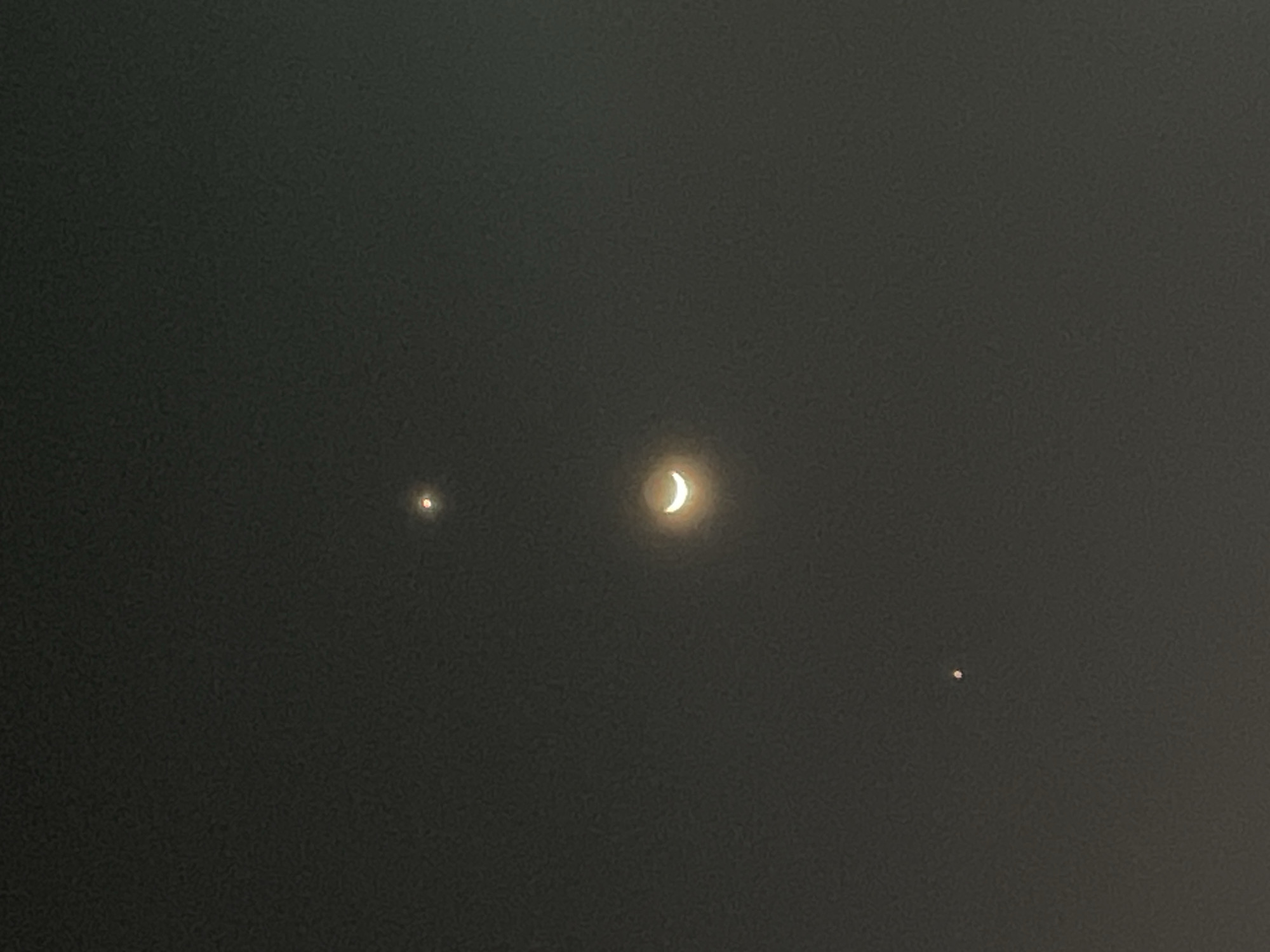This media file depicts a celestial scene in the night sky. Three luminous objects are visible against a dark, overcast backdrop, suggesting a cloudy or hazy night. The most prominent object is a crescent moon, positioned centrally and emitting a bright, soft glow. To its left, a smaller, more point-like light source glows with a reddish hue. To the right of the moon, and slightly lower, is another tiny, distinct point of light, this one appearing white.
The overall impression is one of a quiet, atmospheric night sky. There are no visible people, objects, or specific location cues beyond the suggestion of a night sky, potentially observed from Dar es Salaam, Tanzania. The activity taking place is simply the passive observation of celestial bodies. Notable details include the phase of the moon, the relative positions and apparent brightness of the three celestial objects, and the dim, diffused light that hints at atmospheric conditions. There is no visible text in the image.