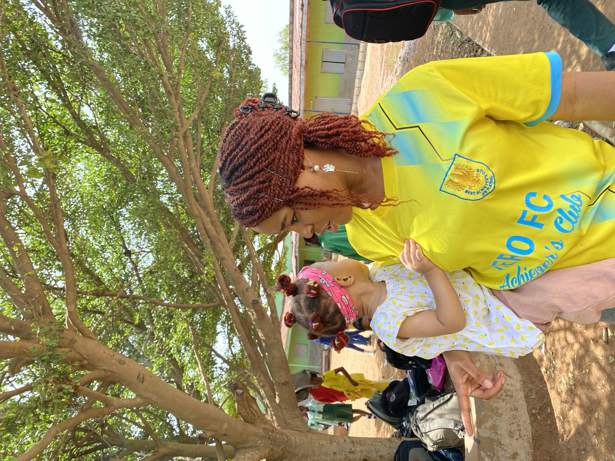 A woman is holding a young child in an outdoor environment in Gura Top, Nigeria. The woman is wearing a yellow soccer jersey with blue accents. The child is wearing a white dress with yellow lemon patterns and a pink headband. Both appear to be standing outside a school. Other people and backpacks can be seen in the background. A large tree is present on the left side of the image.