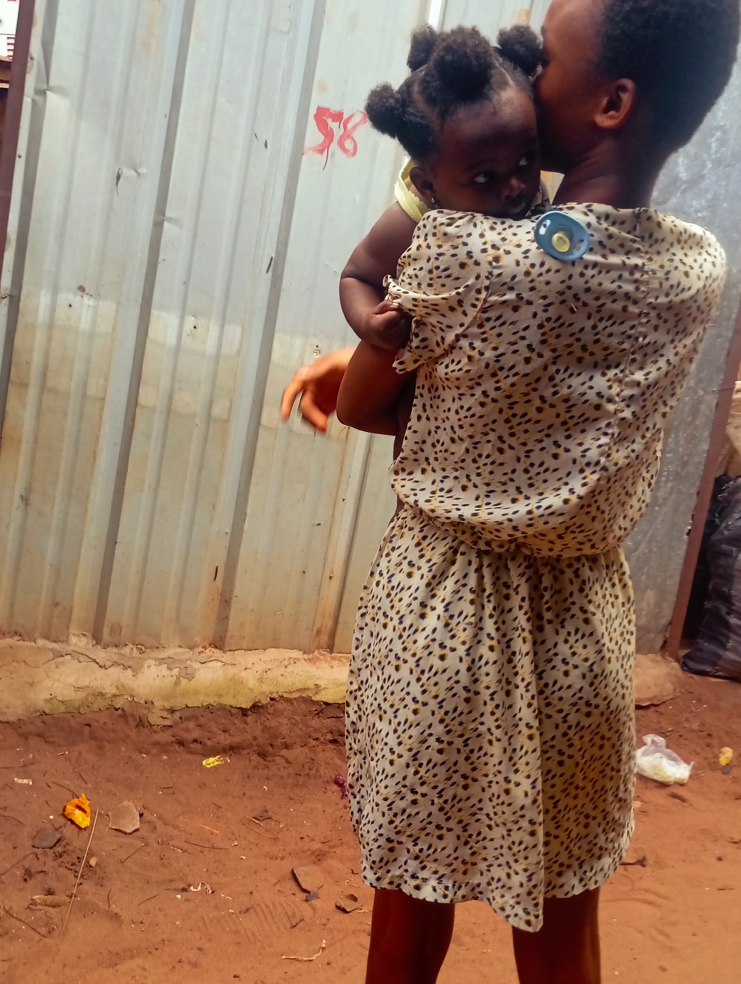 This media file depicts a person holding a baby. The person appears to be a young woman, seen from the back and side, holding the baby up to her cheek in a gesture of affection. The baby is facing away from the camera and is also partially obscured. Both individuals have dark skin tones. The woman is wearing a light-colored dress with a speckled or animal print pattern. A blue pacifier with a yellow ring is attached to the back of her dress.
The setting appears to be outdoors, with a corrugated metal wall forming the background. The ground is made of reddish-brown dirt with some scattered debris, including a piece of orange material, rocks, and what looks like plastic waste. The lighting suggests it could be daytime, possibly overcast. There is a faint red marking on the metal wall that resembles the number "58". The overall impression is of a casual, perhaps domestic or everyday scene. No specific emotion is explicitly conveyed beyond the affectionate gesture between the two subjects.