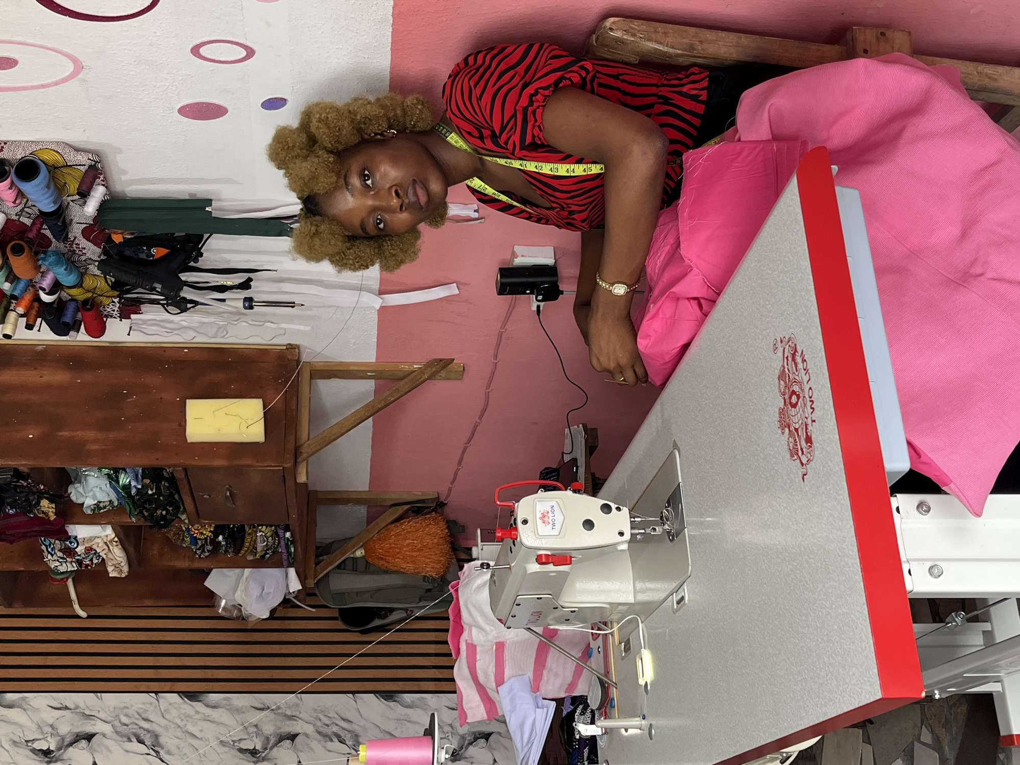 An African woman is seated at a sewing machine in what appears to be a small sewing studio in Badagry, Nigeria. The walls are painted pink and white with decorative circles. The woman has light brown curly hair, a zebra-striped red and black top, and a yellow measuring tape around her neck. She is looking directly at the camera with a neutral expression. She has a watch on her left wrist.
The sewing machine is white with red accents and is surrounded by pink fabric. A small wooden shelf holds various sewing supplies, including spools of thread and fabric scraps. The floor is tiled in a marble-like pattern. A broom leans against the wall in the background. The scene is brightly lit. There are electrical outlets and wiring visible on the wall behind the woman.
