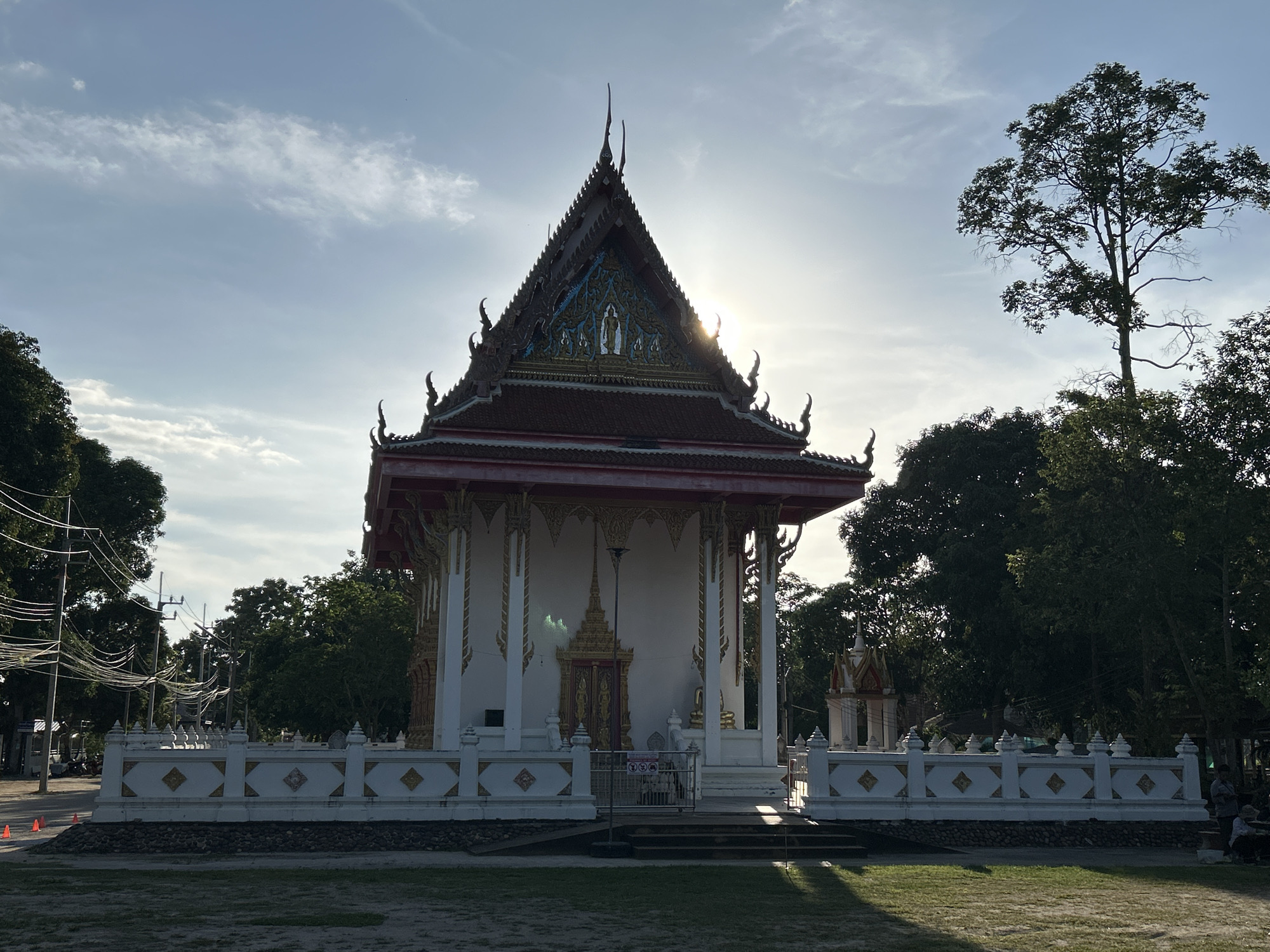 The image shows a Thai Buddhist temple in ตำบลเกาะพะงัน, Thailand. The temple is a white building with a red and gold roof, surrounded by a white fence. The sun is shining behind the temple, creating a halo effect. There are trees around the temple and a grassy area in front of it. The sky is blue with some clouds.