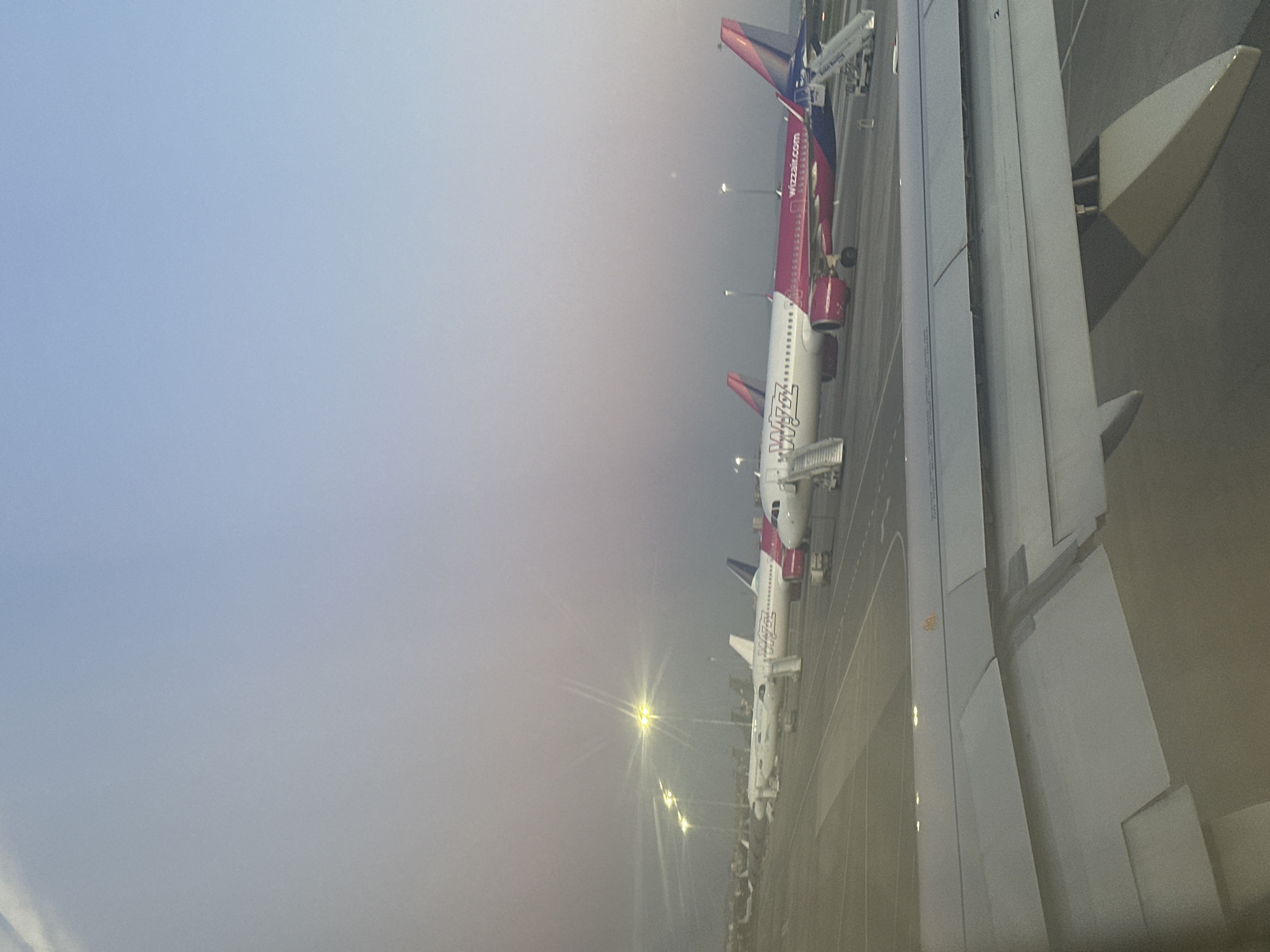 The image shows an aerial view of a row of airplanes, predominantly Wizz Air, parked on a tarmac, likely at Sofia Airport in Sofia, Bulgaria. An airplane wing is visible in the foreground. The atmosphere appears foggy or hazy, and the sky is light. Streetlights illuminate the area. The airport is quiet, with no visible people or significant activity around the airplanes.