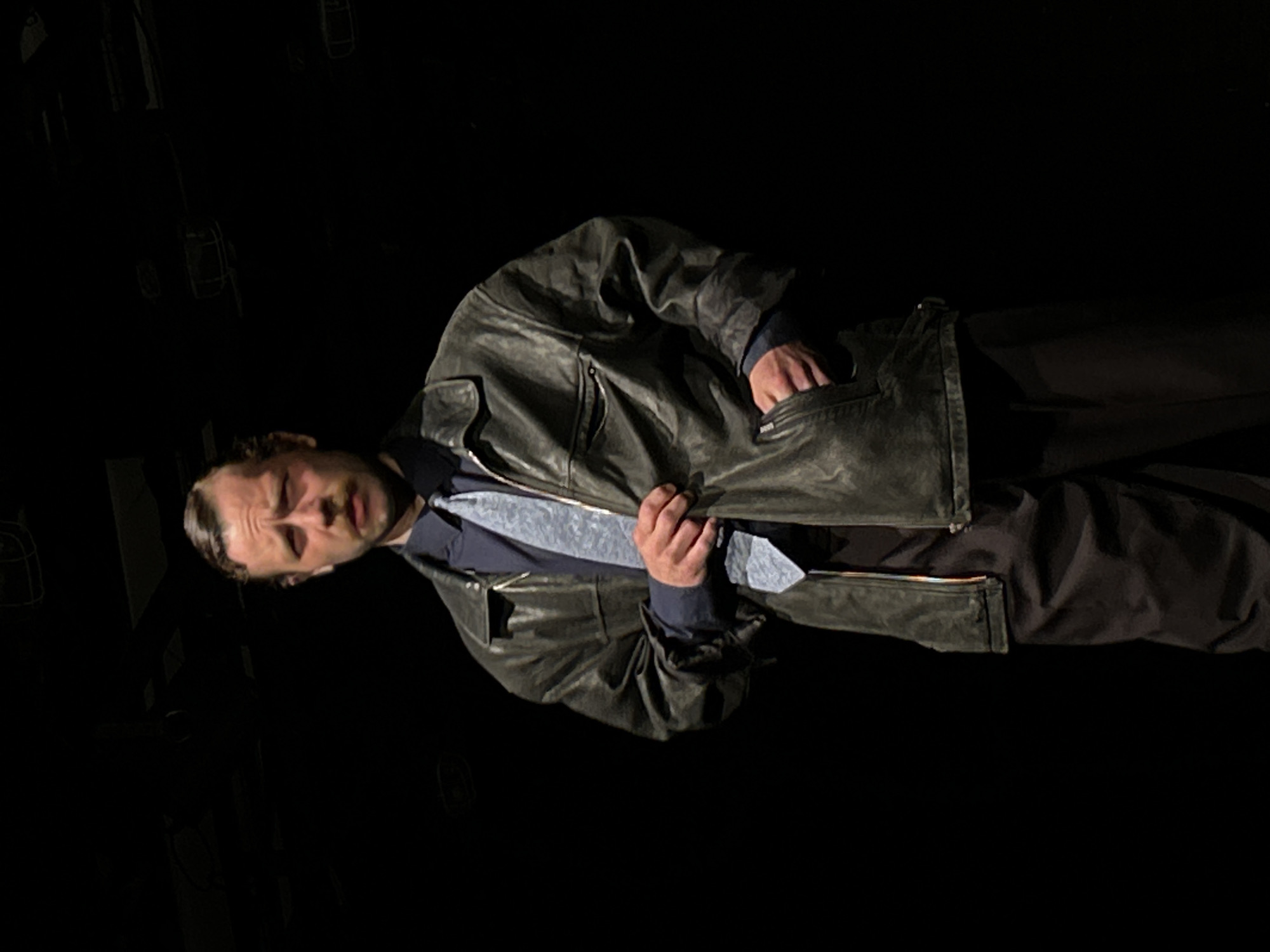 A man is centered in the image, turned at an angle. He is wearing a dark gray leather jacket, a dark shirt, and a light gray tie. His pants are dark as well. The background is entirely black, and the location is unknown. The man's hand is in his jacket pocket.